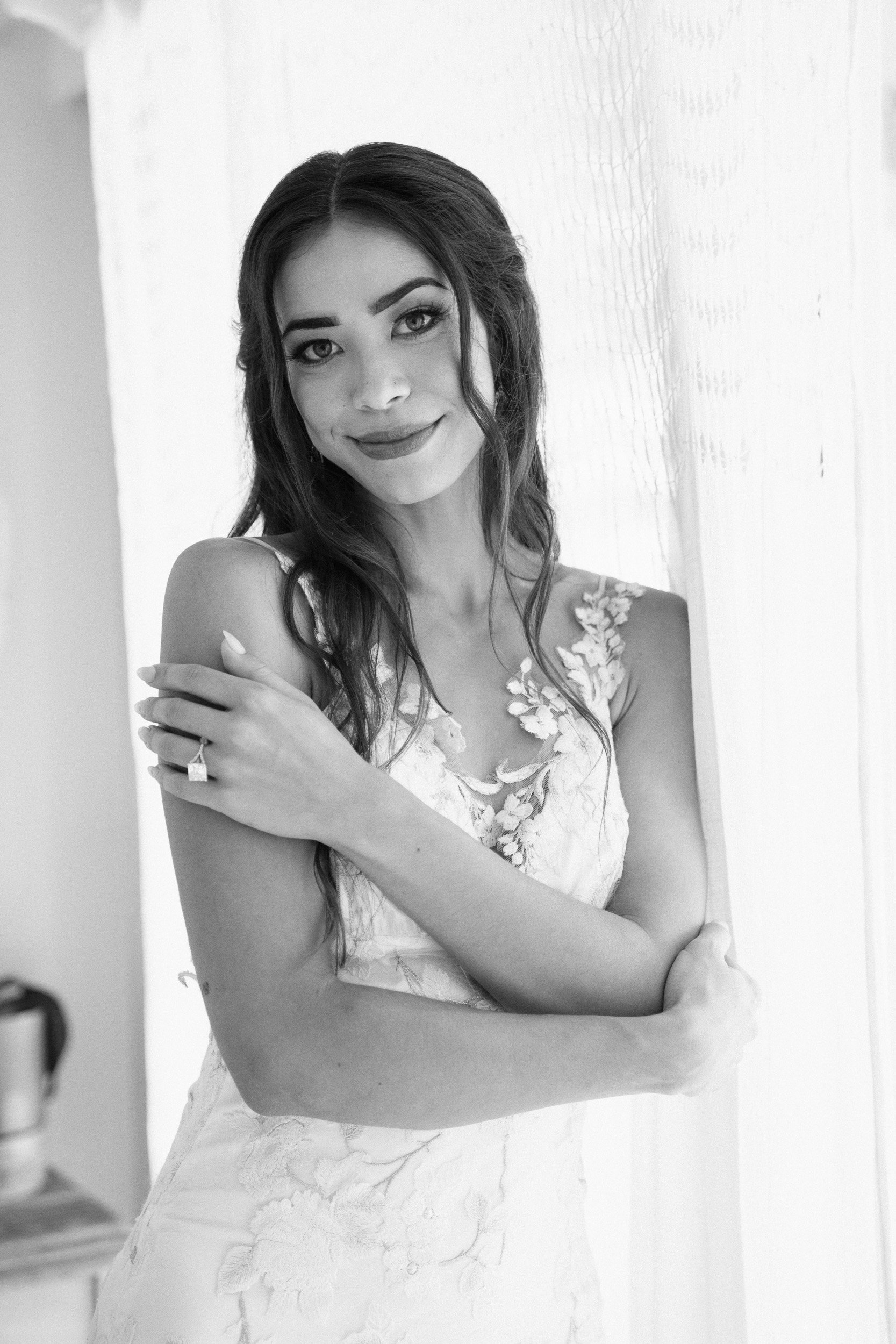 Black and white photo of a woman with long wavy hair, wearing a dress with floral embroidery, smiling and looking at the camera, standing near a textured wall.