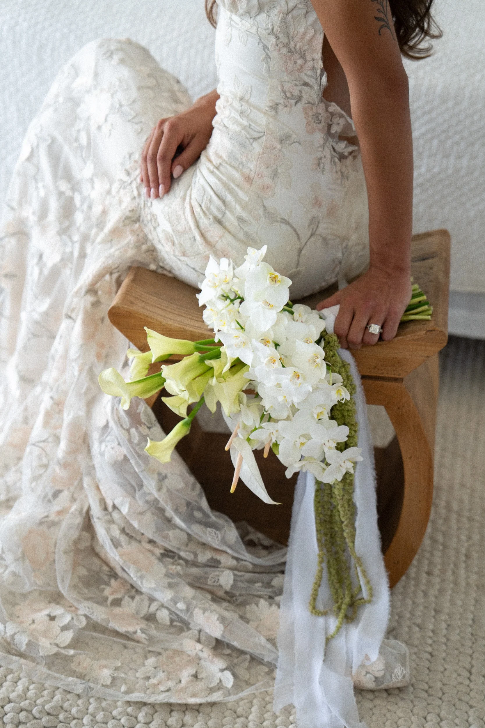Close-up of a woman in a wedding dress sitting on a wooden bench, holding a bouquet of white orchids, with lace fabric and decorative elements visible.