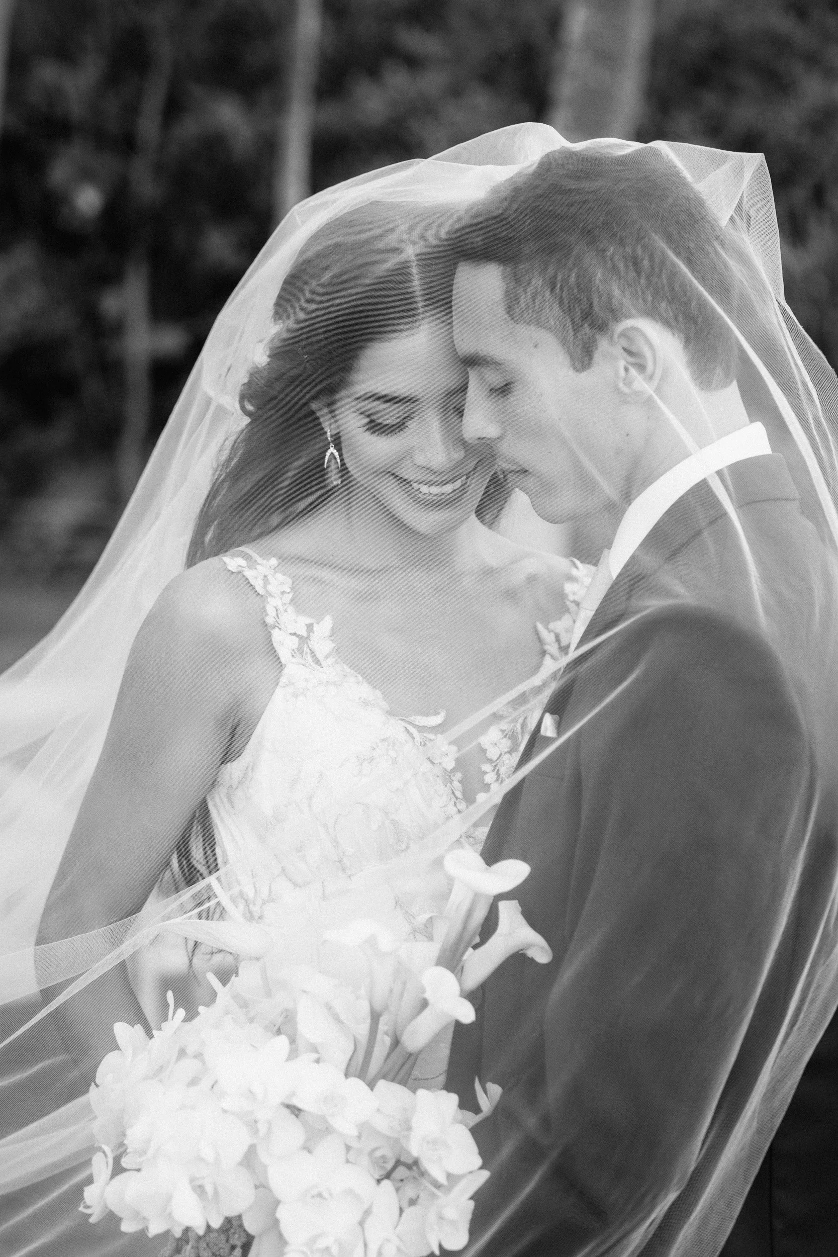 A bride and groom with their foreheads touching under a wedding veil, smiling with eyes closed, outdoors with trees in the background, black and white photograph.