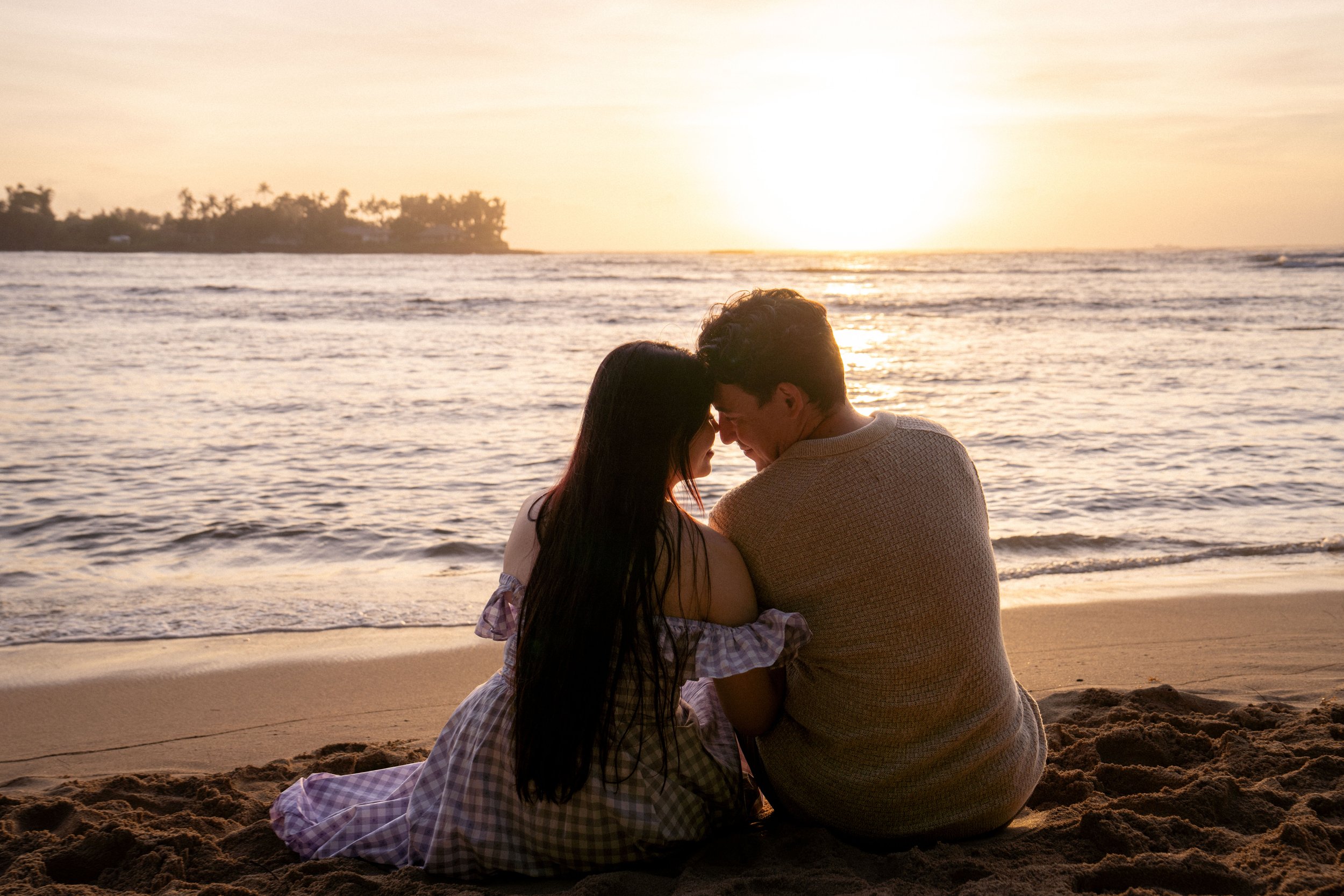 A couple sitting on the beach at sunset, with their foreheads touching and eyes closed, facing each other near the shoreline.