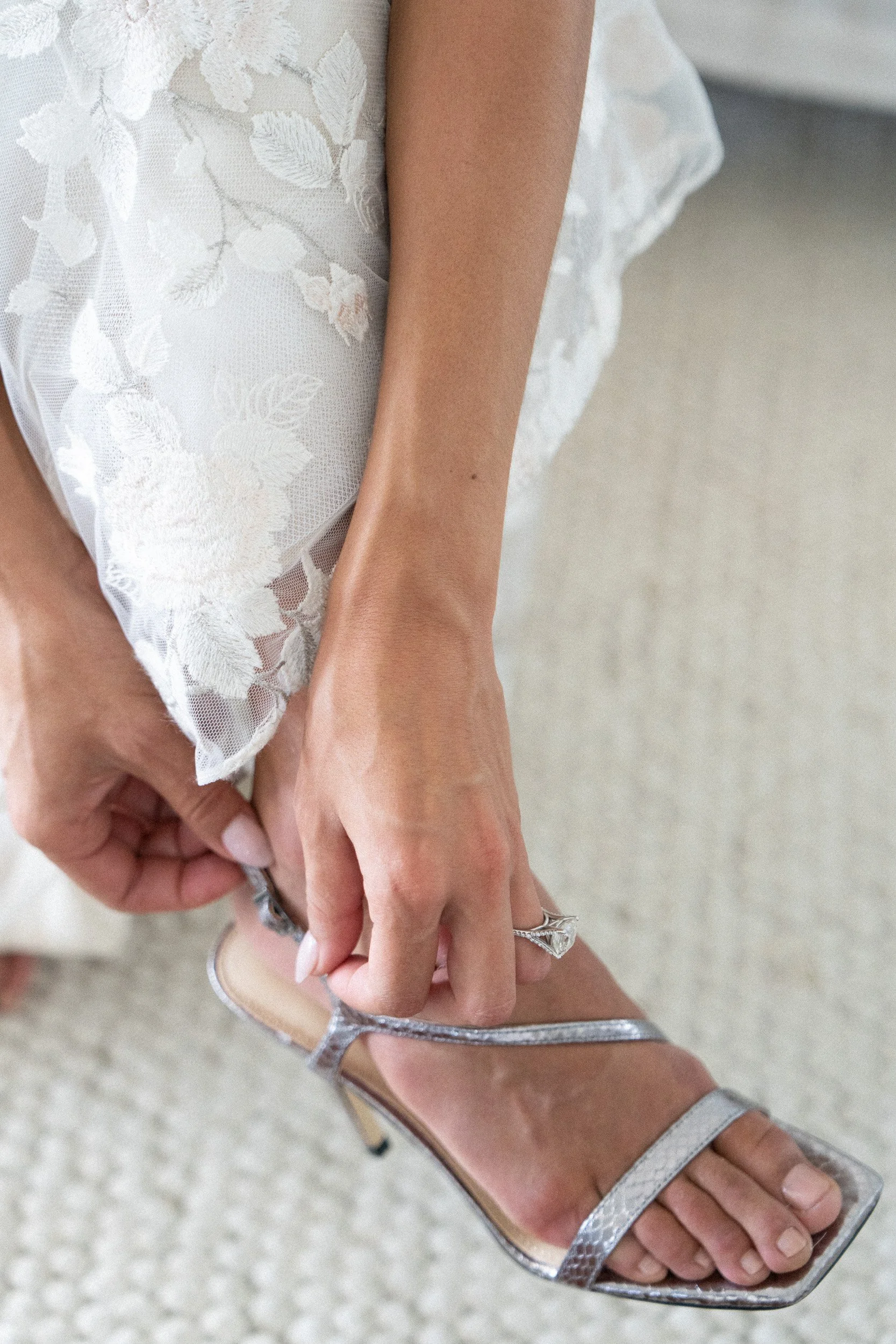 Close-up of a woman’s foot in a high-heeled sandal with metallic straps, being held and adjusted by her hand. She is wearing a delicate, white lace dress.