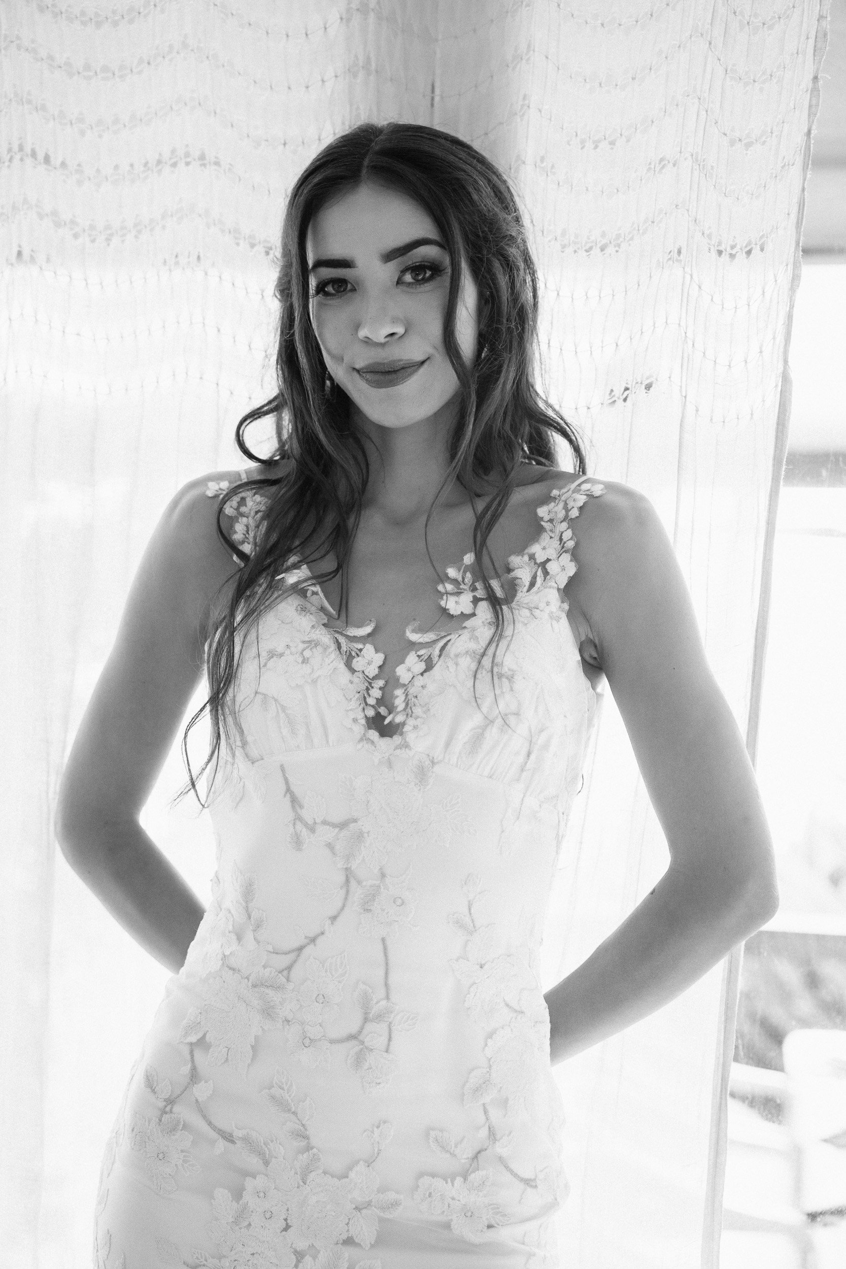 Black and white photo of a woman in a white dress with floral embroidery, standing with hands on hips, smiling softly, in front of a lace curtain and a window.