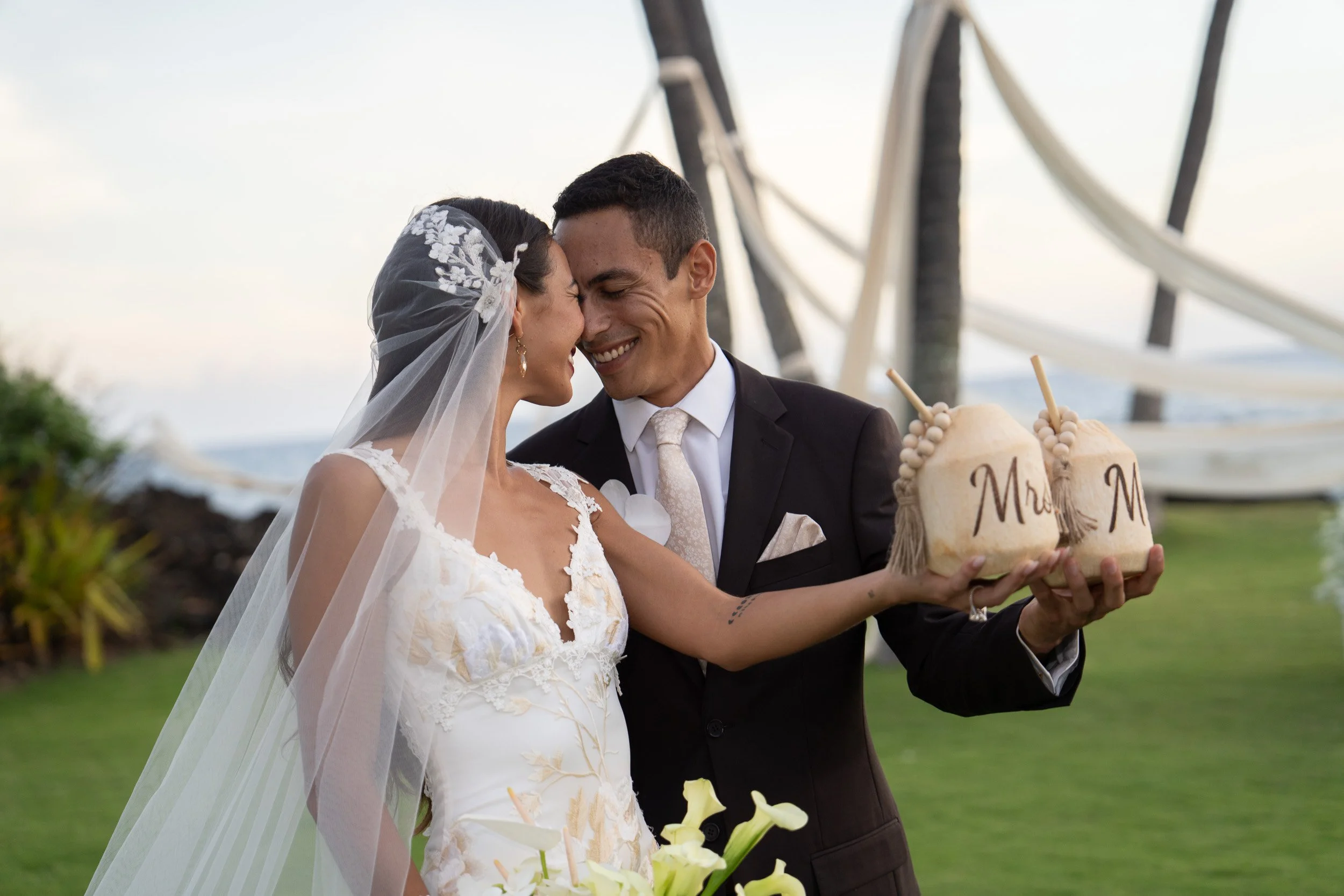 A bride and groom smiling and touching foreheads at their wedding outdoors, with a large Ferris wheel in the background. The bride is wearing a wedding dress with lace details and a veil, while the groom is in a black suit with a white tie. They are 