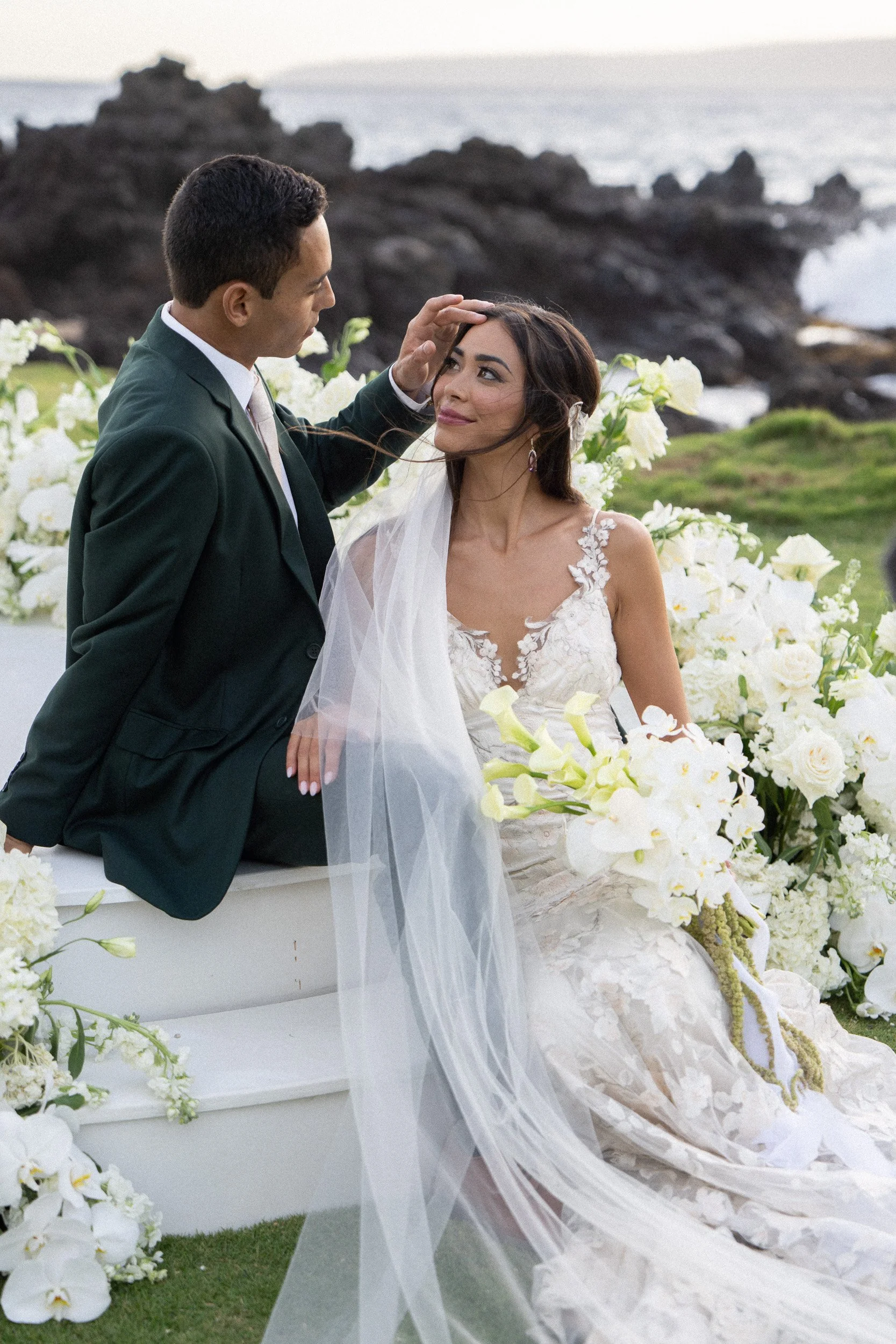 A groom touching a bride's forehead during a wedding ceremony outdoors, with white floral arrangements and rocky shoreline in the background.