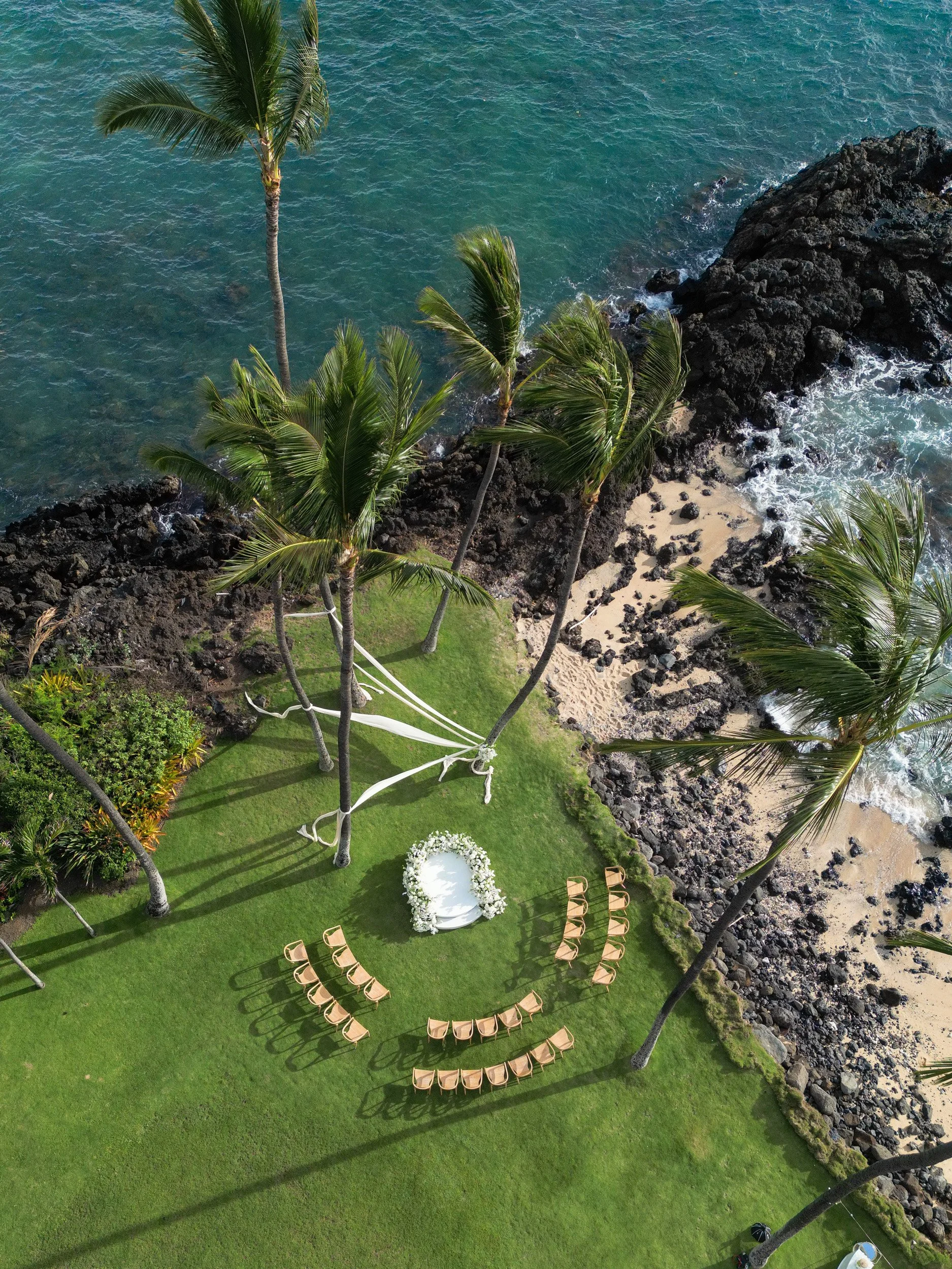Overhead view of a tropical beach wedding setup with chairs arranged in a semi-circle, a floral arch, palm trees, rocky shoreline, and the ocean.