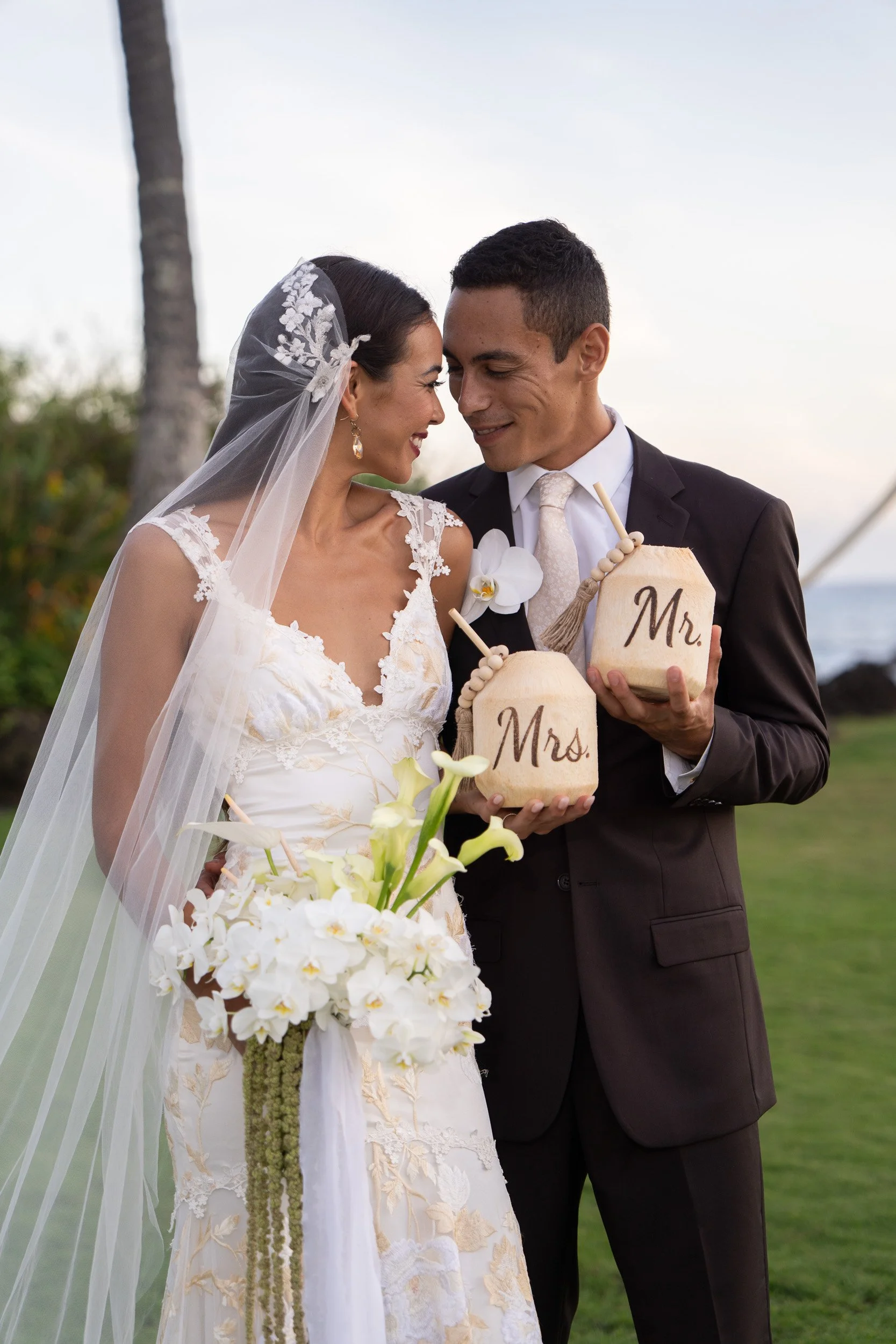 A bride in a lace wedding dress and a veil smiling and leaning towards a groom in a black suit and white tie, holding wooden signs with 'Mr.' and 'Mrs.', with tropical plants and a tree in the background.