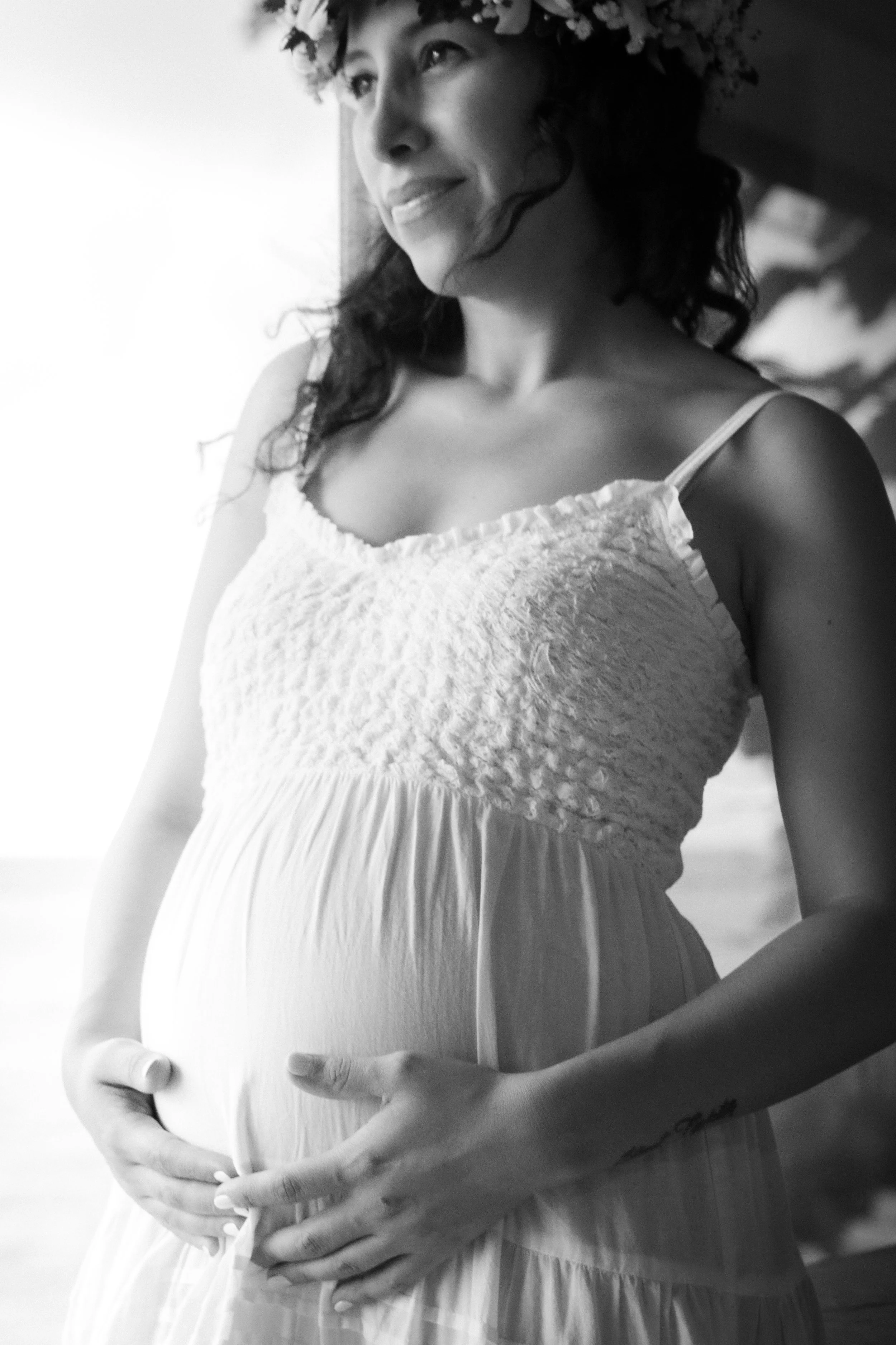 A pregnant woman wearing a flowy, sleeveless dress with ruffled details on the bodice, holding her belly with both hands, and smiling softly. She has dark, curly hair and a flower crown on her head. The image is in black and white.