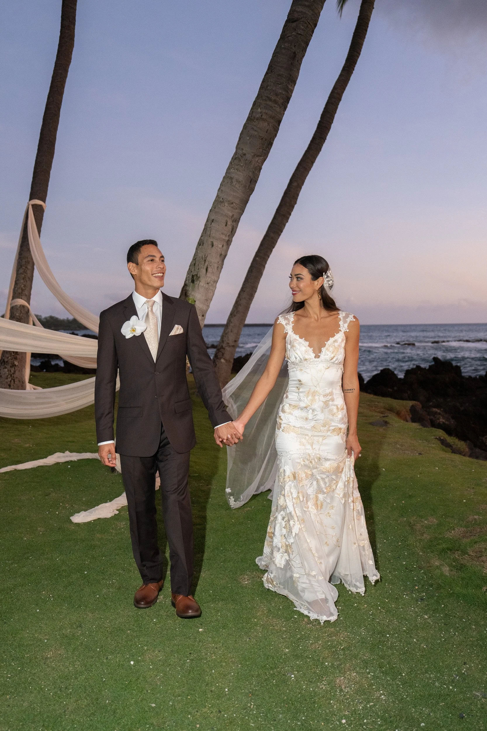 A bride and groom holding hands during their wedding ceremony on a grassy area at the beach during sunset, with palm trees and the ocean in the background.