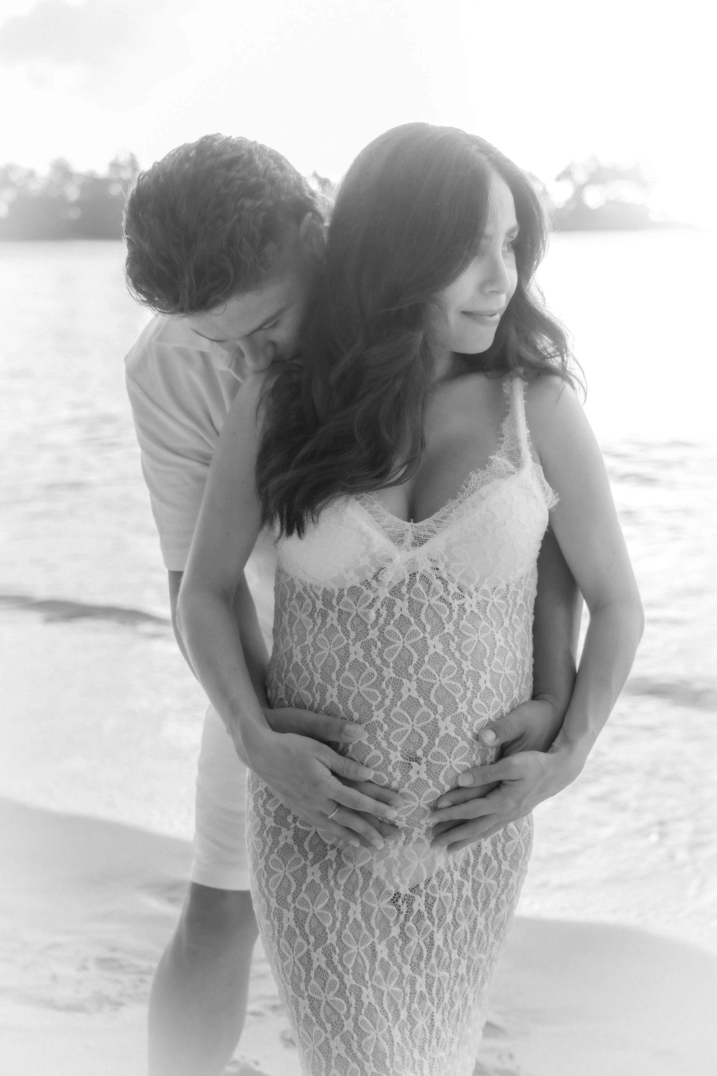 A couple on the beach in oahu during a photoshoot by isis monteux, with the man embracing the pregnant woman from behind, both smiling softly, in black and white.