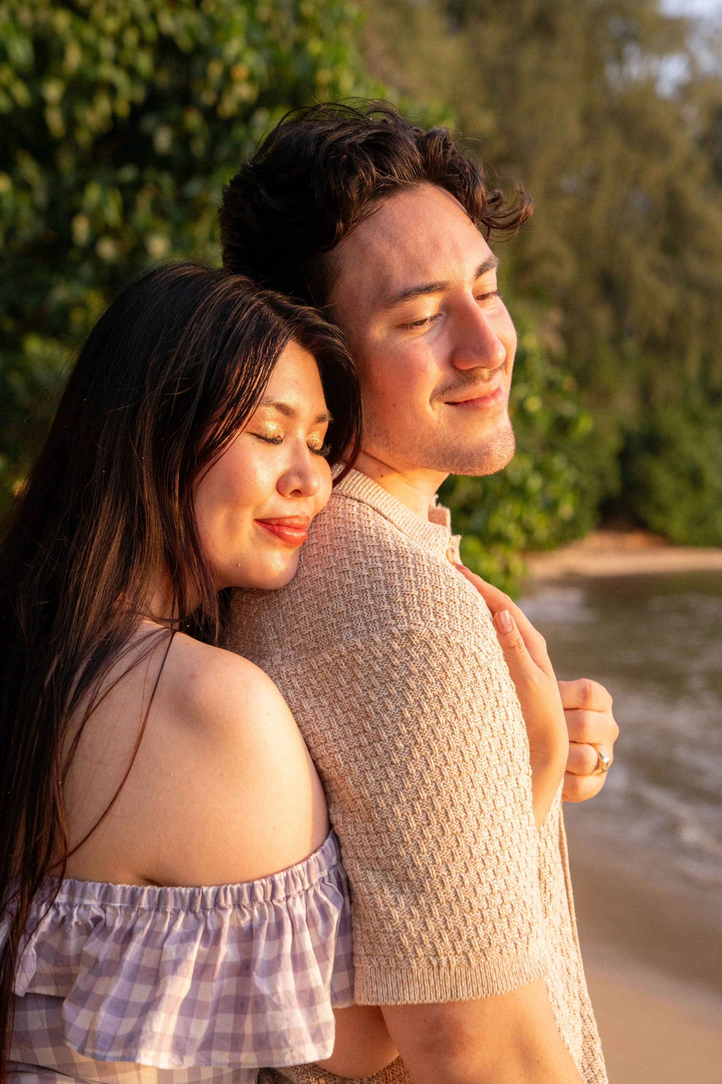 A woman and a man embrace on a beach during sunset, both with closed eyes, smiling gently, with lush green trees in the background.