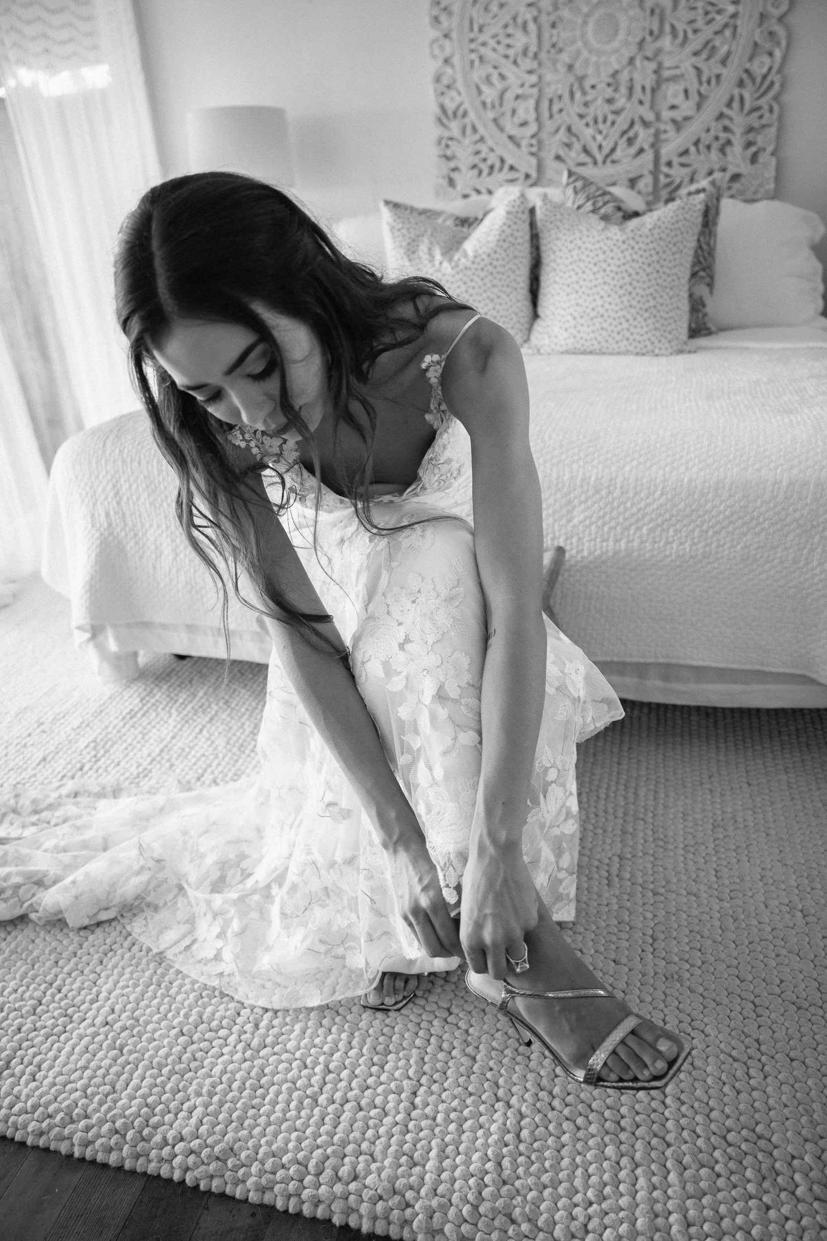 A woman in a lace dress sitting on a bed, adjusting her high heels, with pillows and an ornate wall hanging in the background.