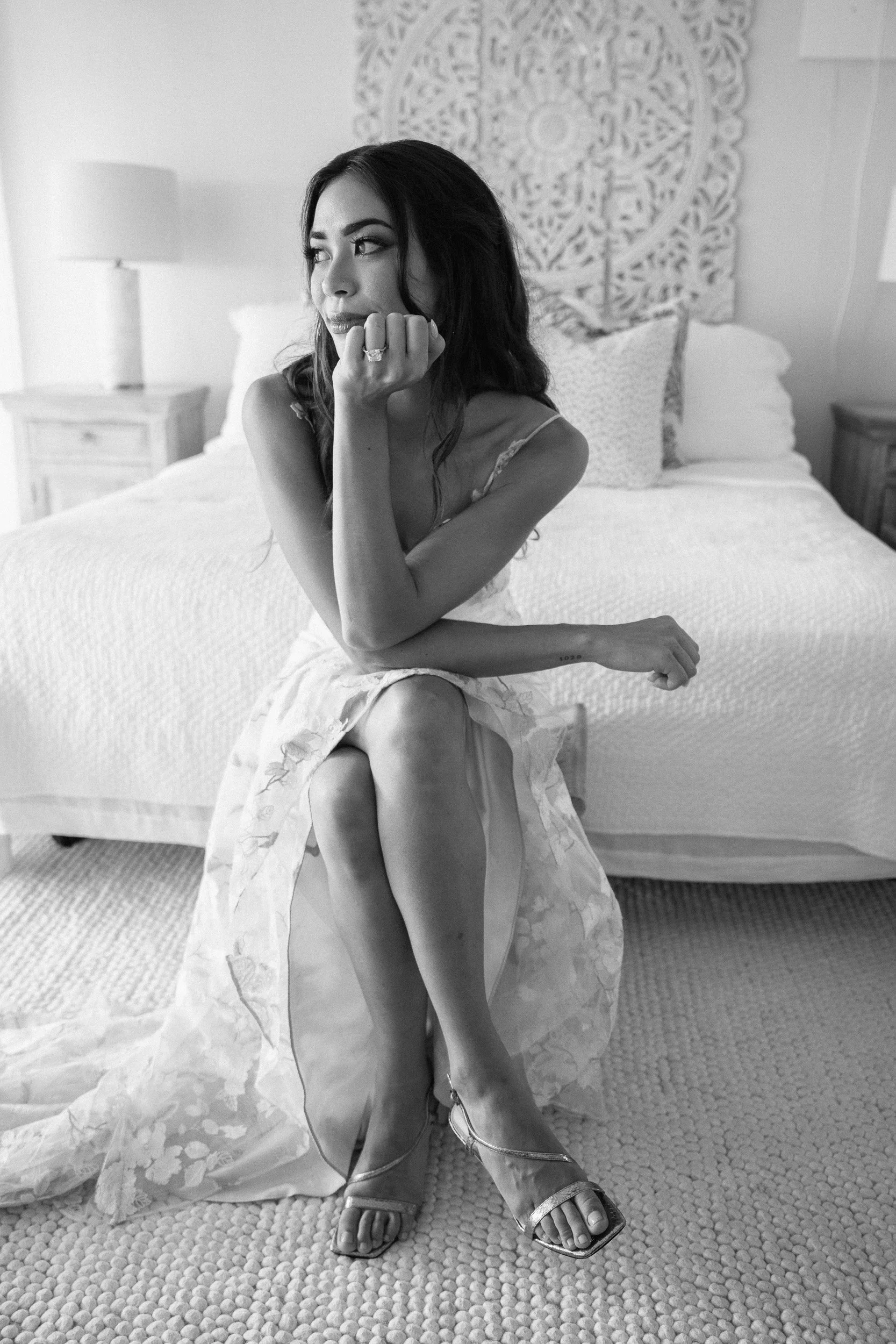 A woman sitting on a bed, resting her chin on her hand, wearing a dress and high-heeled sandals, with a decorative wall hanging behind her in a bedroom.
