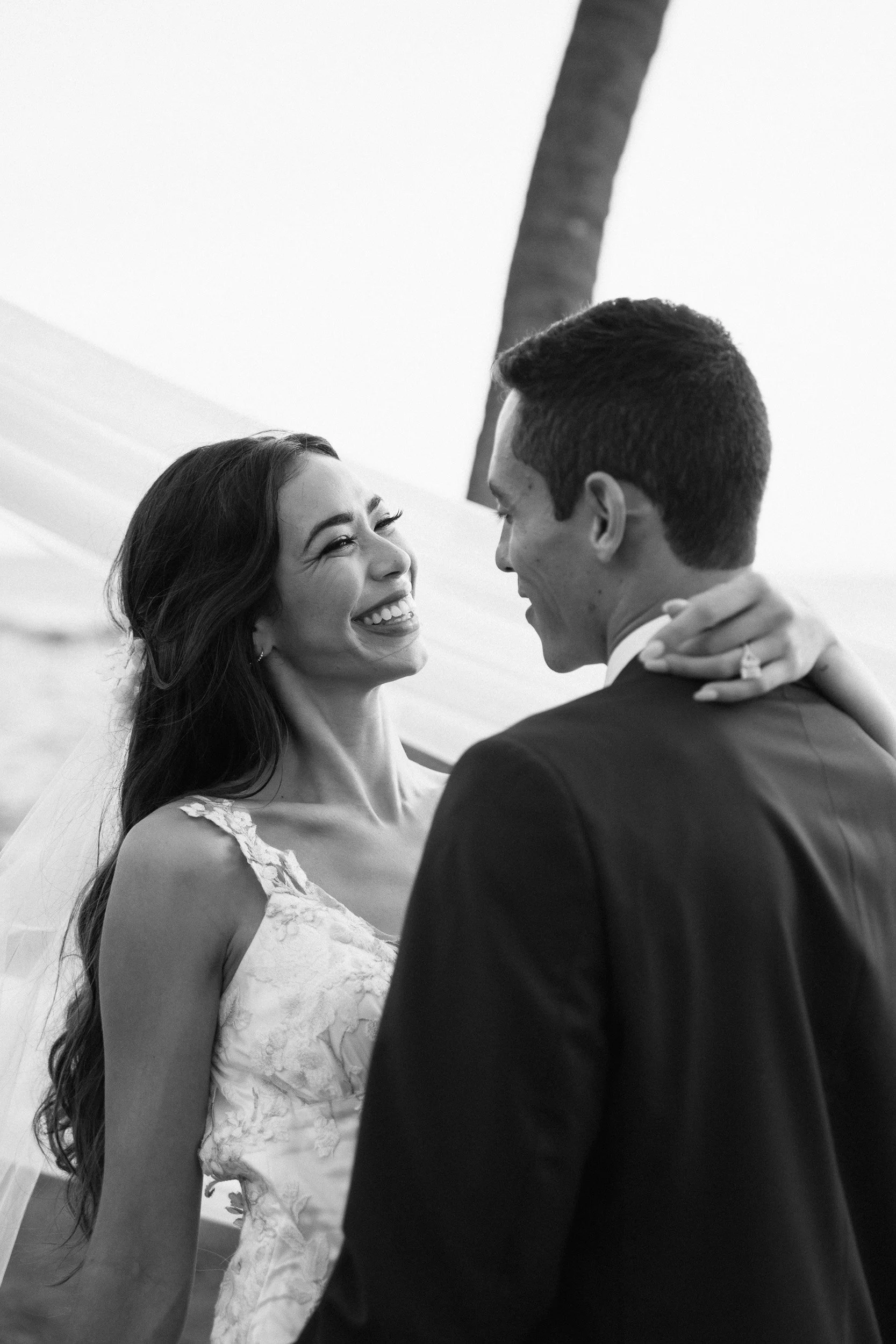 A smiling woman in a wedding dress looks at a man in a suit, who is smiling back at her, while they are dancing outdoors with a palm tree in the background.