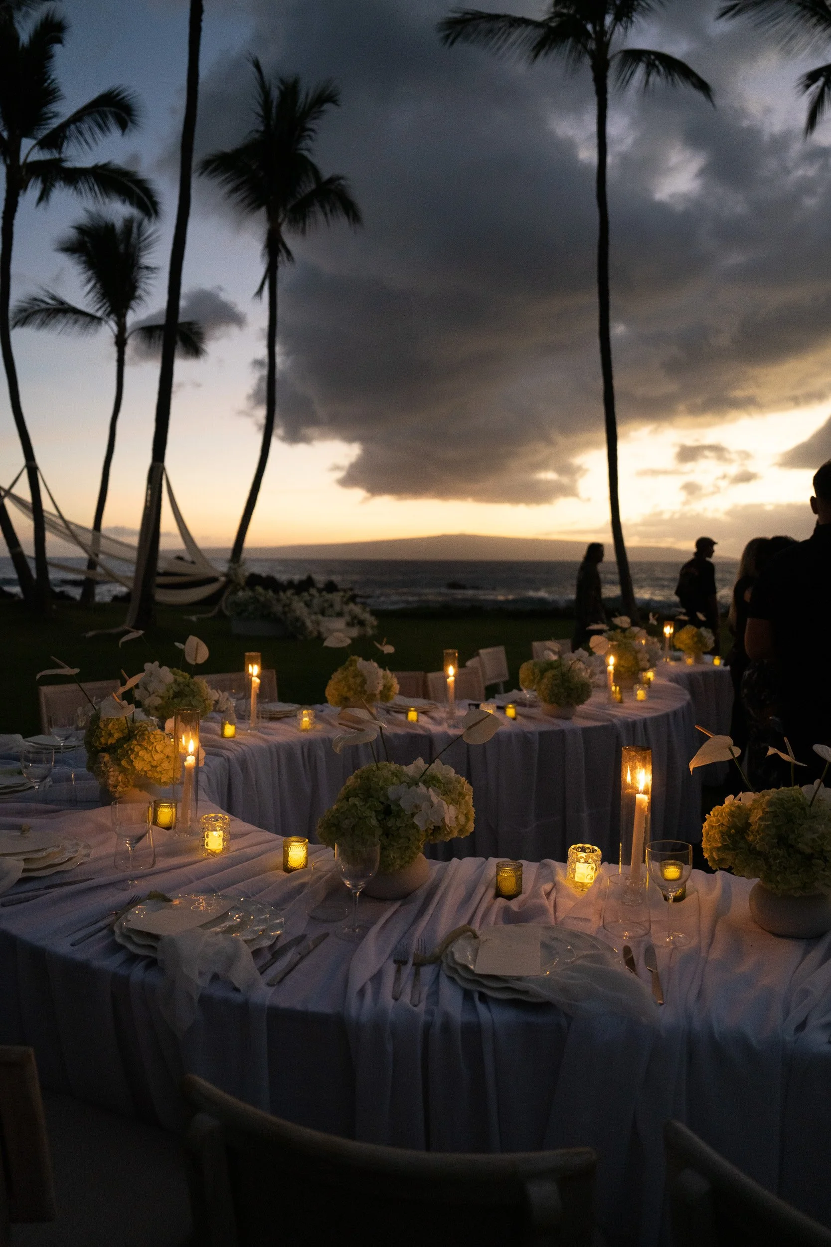 An outdoor dinner setup by the beach with white tablecloths, floral centerpieces, and candles. There are several tall palm trees and a cloudy sky at sunset or dusk.
