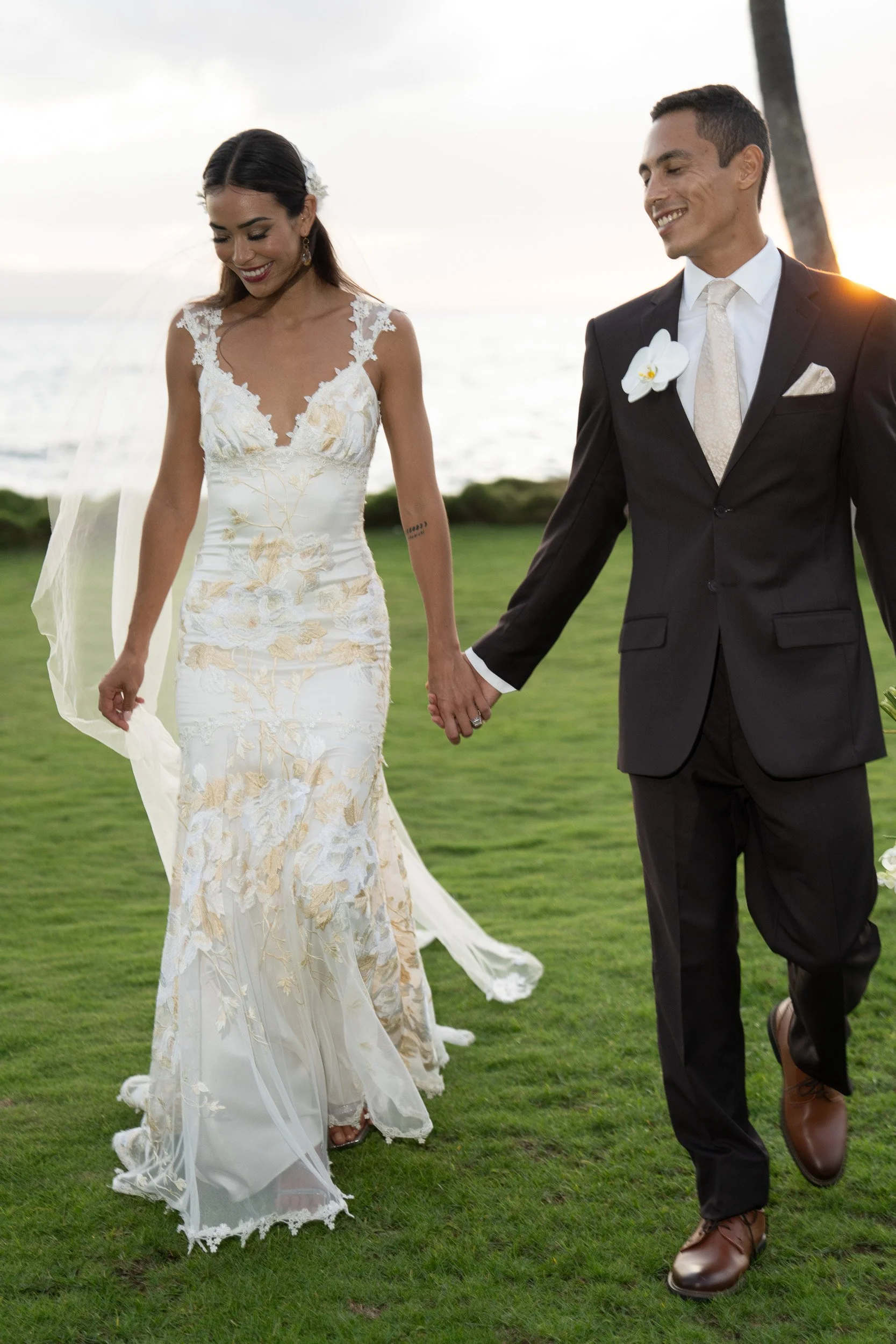 A bride in a white wedding dress with gold embroidery and a groom in a black suit with a white shirt and tie, holding hands and walking on grass near the ocean at sunset.