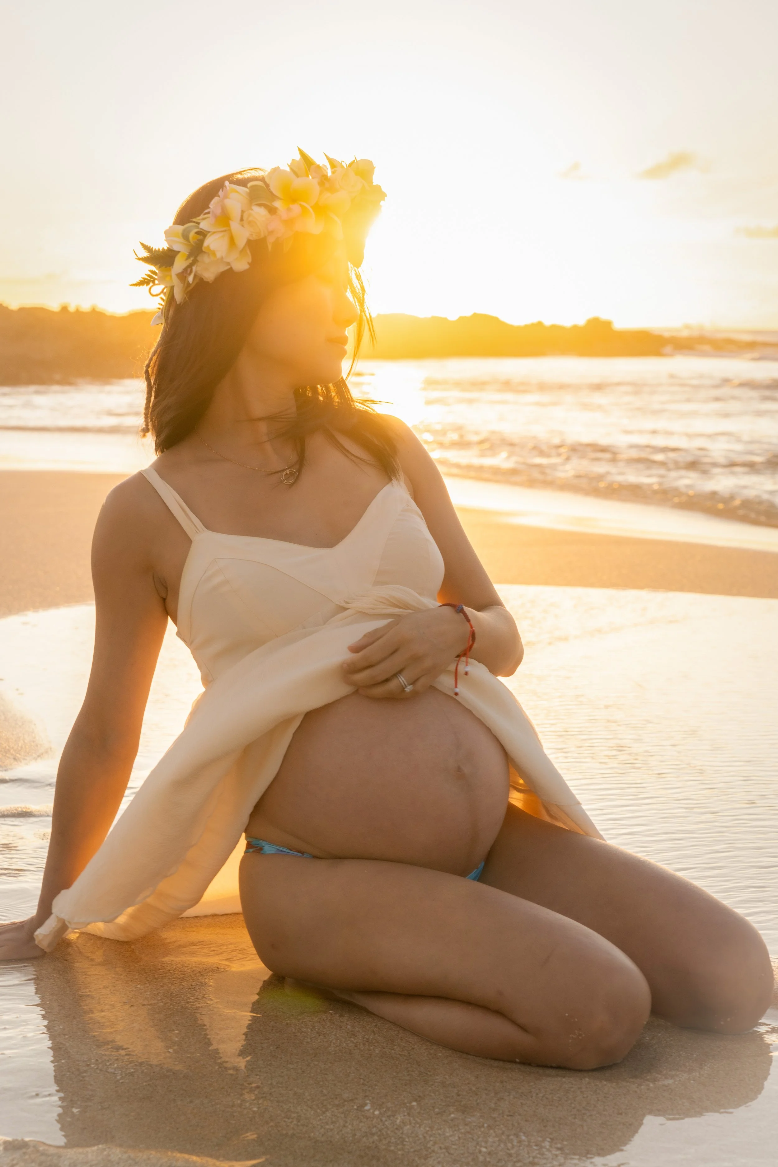 Pregnant woman sitting on the beach at reiki beach, north shore oahu, during sunset, wearing a flower crown and a white dress.