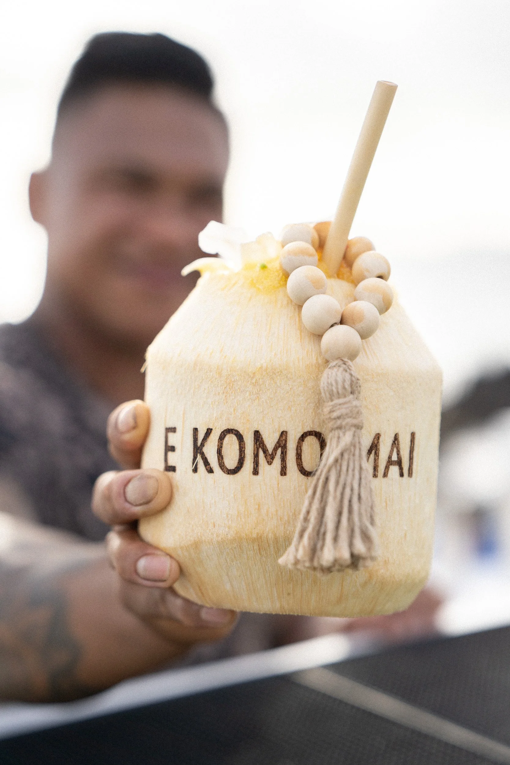 Person holding a wooden coconut beverage container with a white straw and decorative beads with a tassel, with a blurred background.
