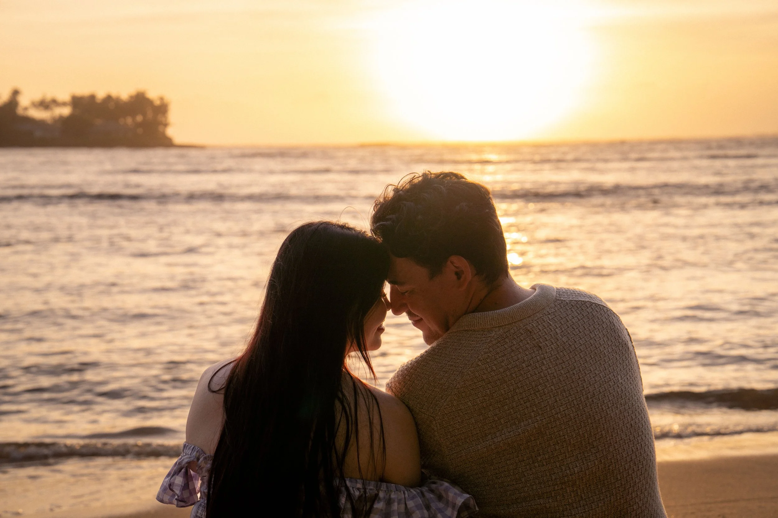 A couple sharing a romantic moment touching foreheads and smiling at the beach during sunset, with calm ocean waves and a distant landmass in the background.