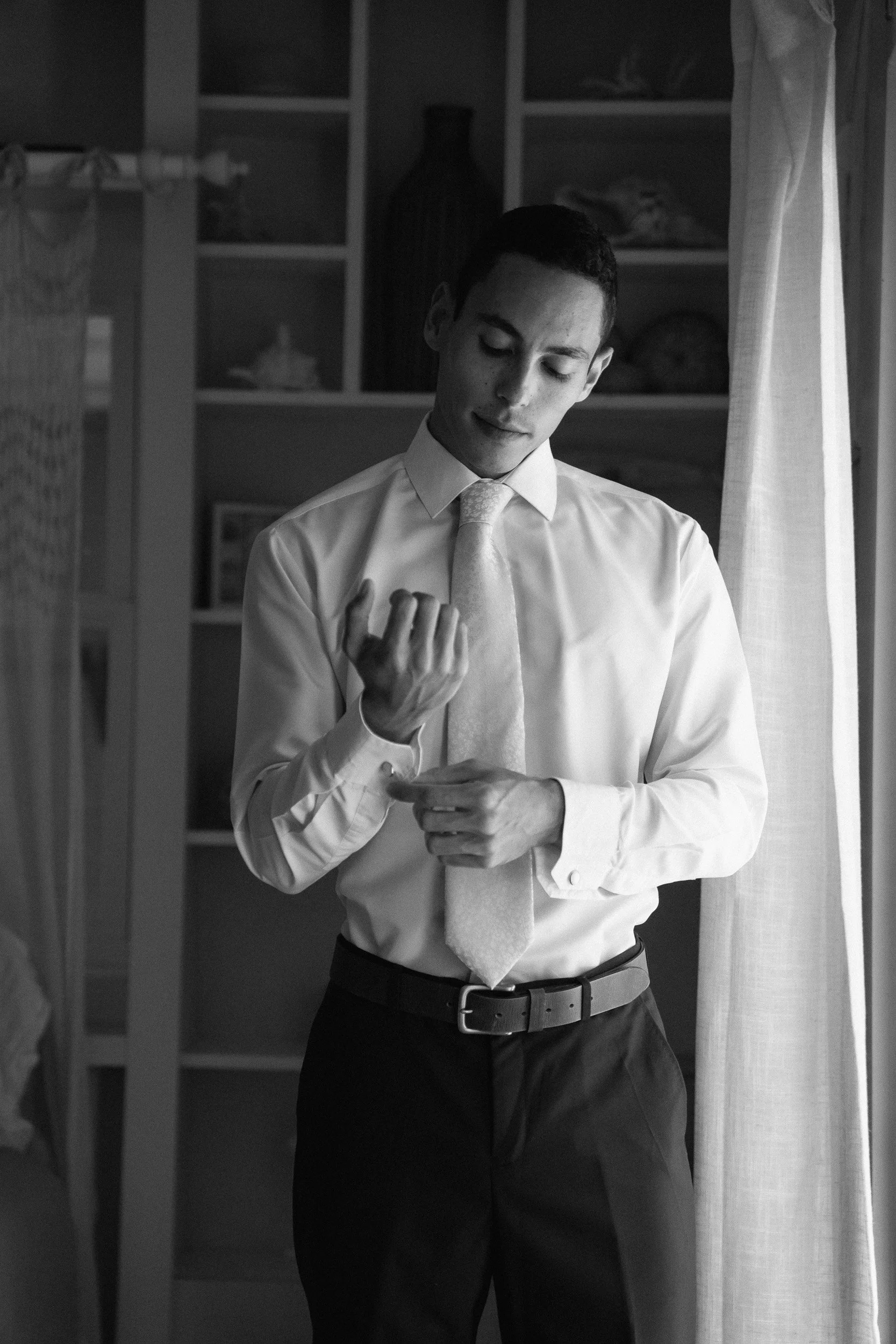 A young man in a dress shirt and tie is adjusting his tie in front of a window with curtains, with a bookshelf in the background.