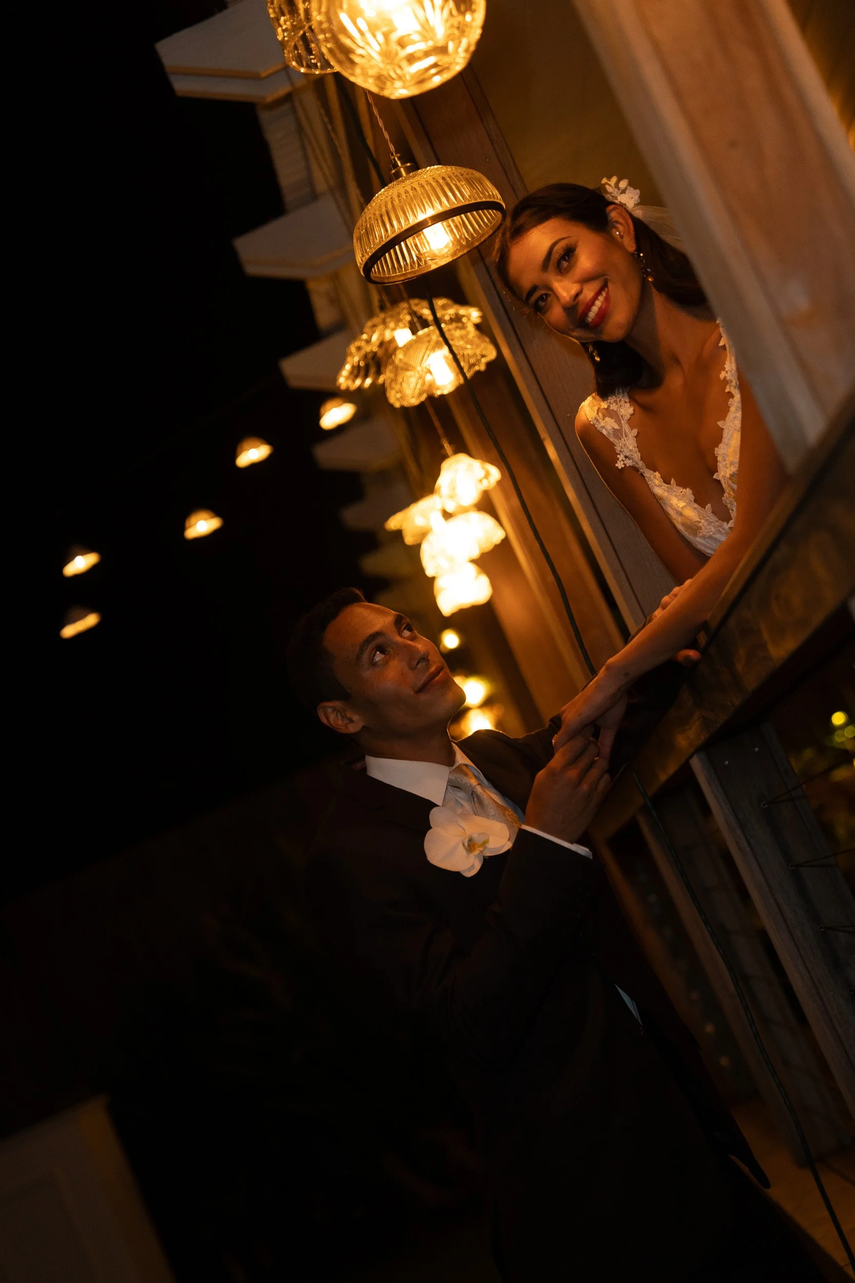 A smiling woman in a white lace dress peeks over a wooden surface, while a man in a tuxedo sits beside her, with warm hanging lights illuminating the scene in a cozy indoor setting.