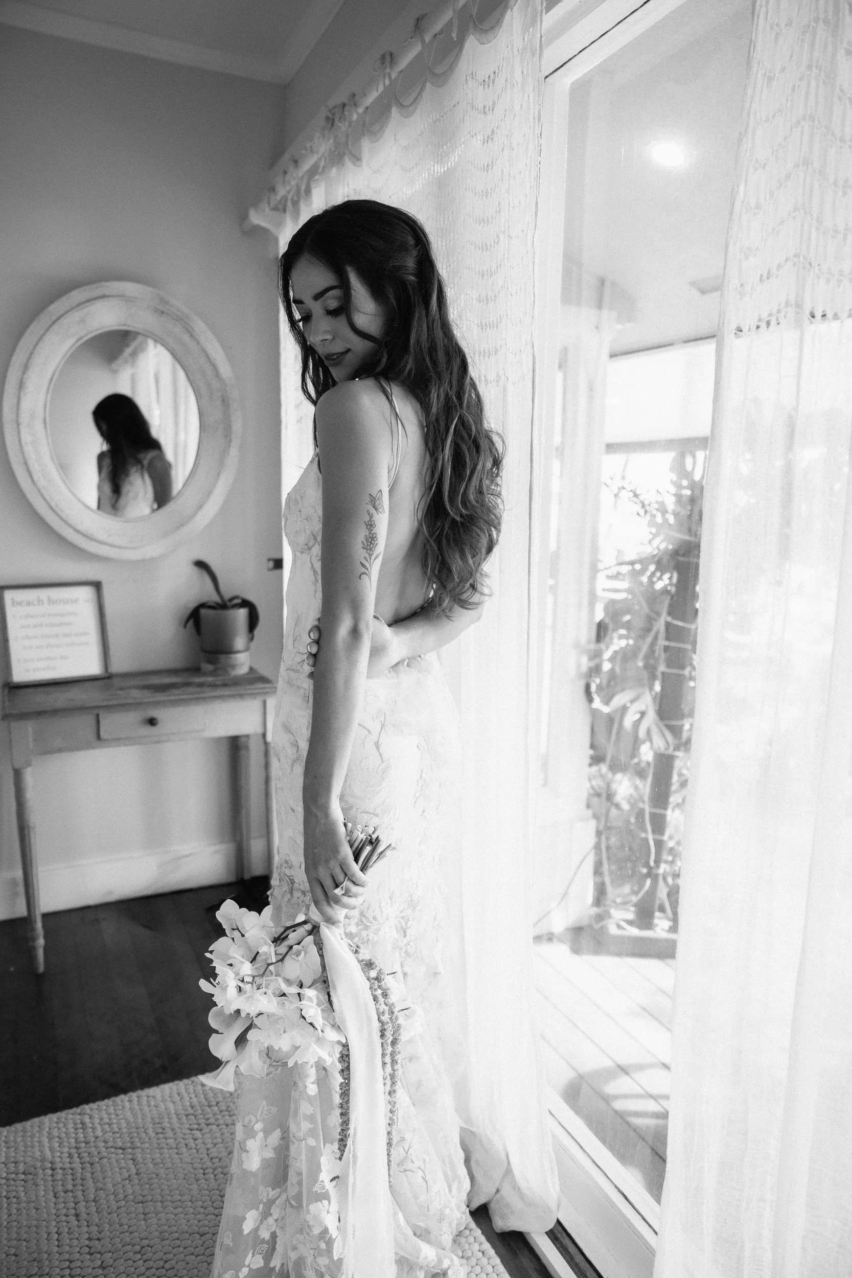 Black and white photo of a woman in a wedding dress, holding a bouquet, standing by a window with sheer curtains, with a mirror reflecting her face.