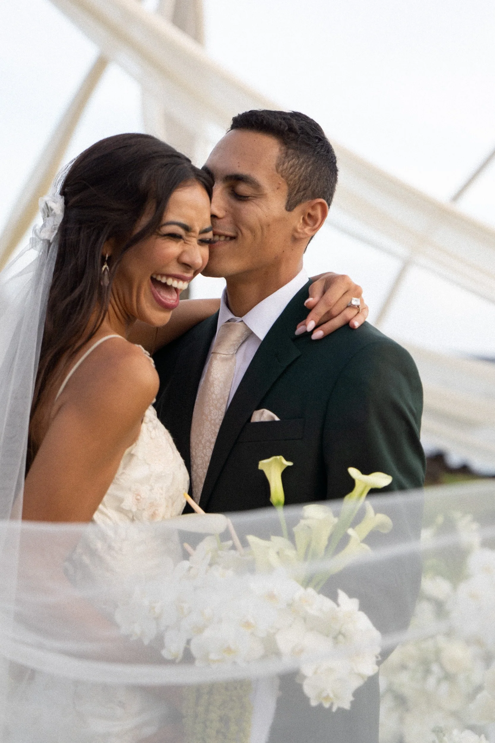 A bride and groom celebrating their wedding, smiling and embracing each other while standing behind a floral arrangement decorated with white calla lilies.