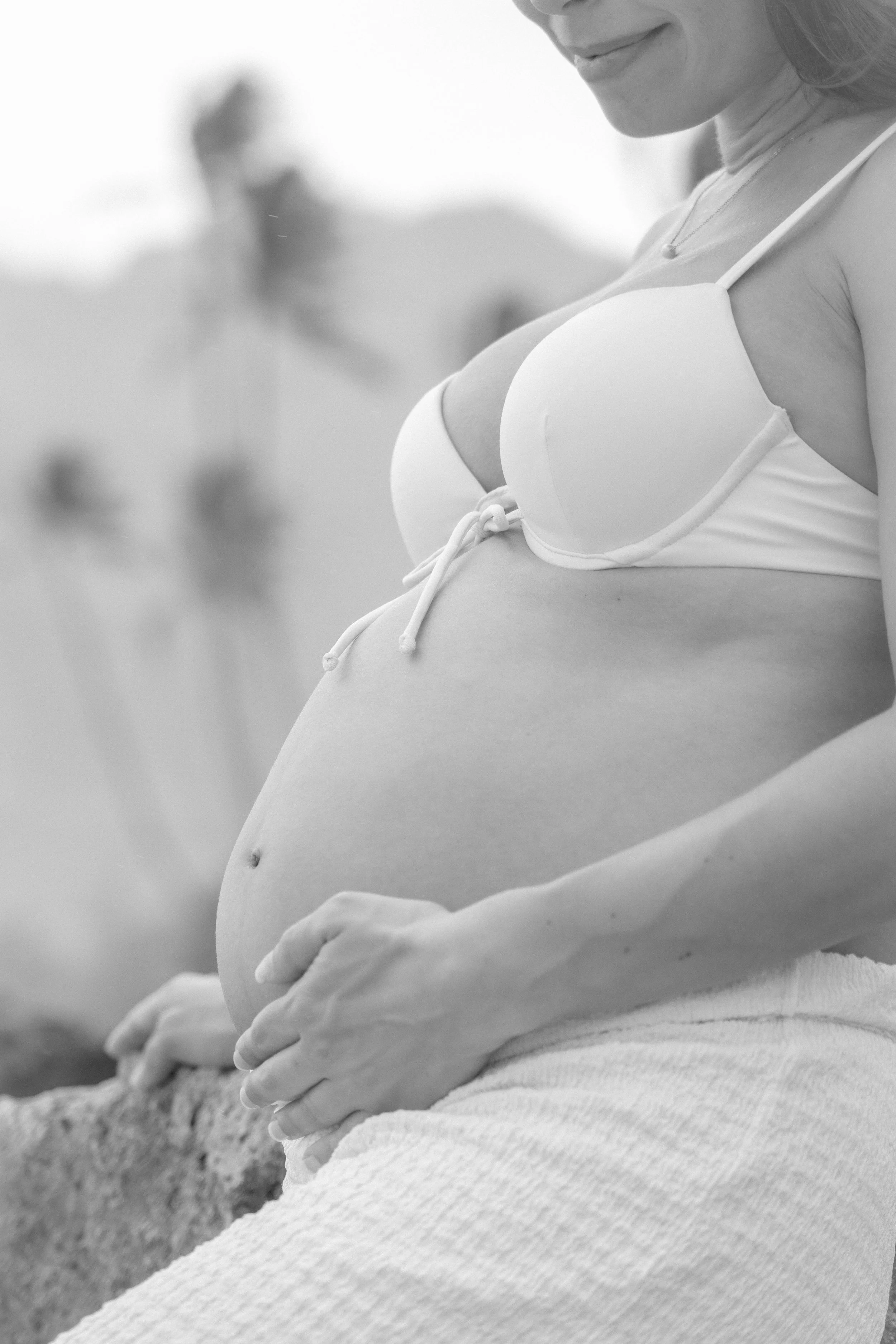 Black and white photograph of a pregnant woman wearing a bikini top, sitting outdoors, with her hands resting on her baby bump.