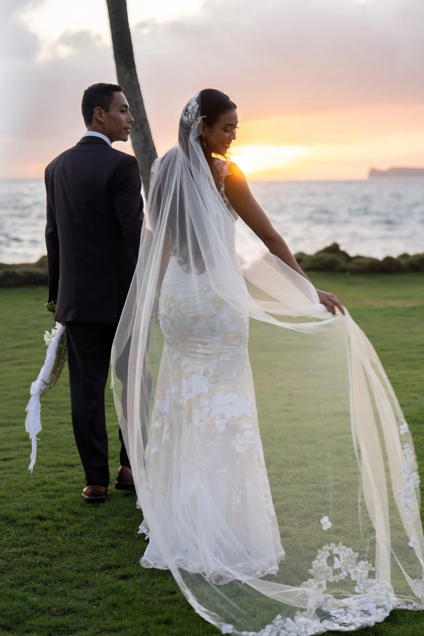 A bride in a white lace wedding dress and veil holding the train of her dress, standing beside a groom in a black suit, with a sunset over the ocean and a palm tree in the background.
