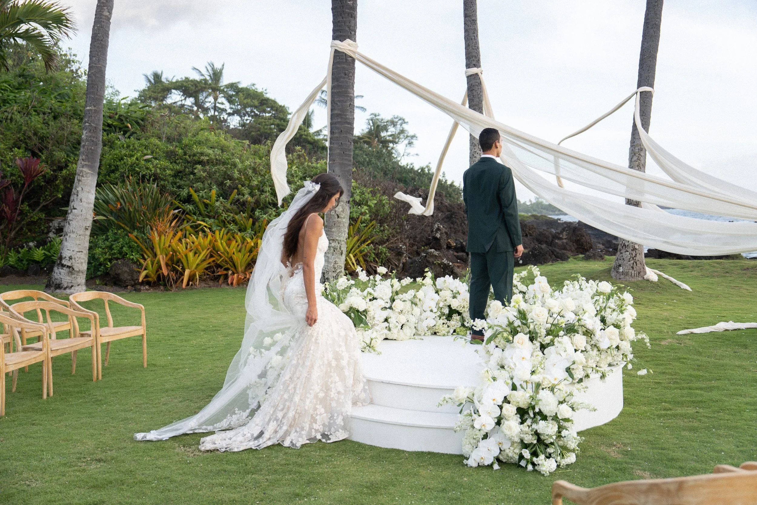 A bride and groom stand at an outdoor wedding ceremony altar decorated with white flowers and fabric, near palm trees and lush greenery, with wooden chairs arranged on the grassy area.