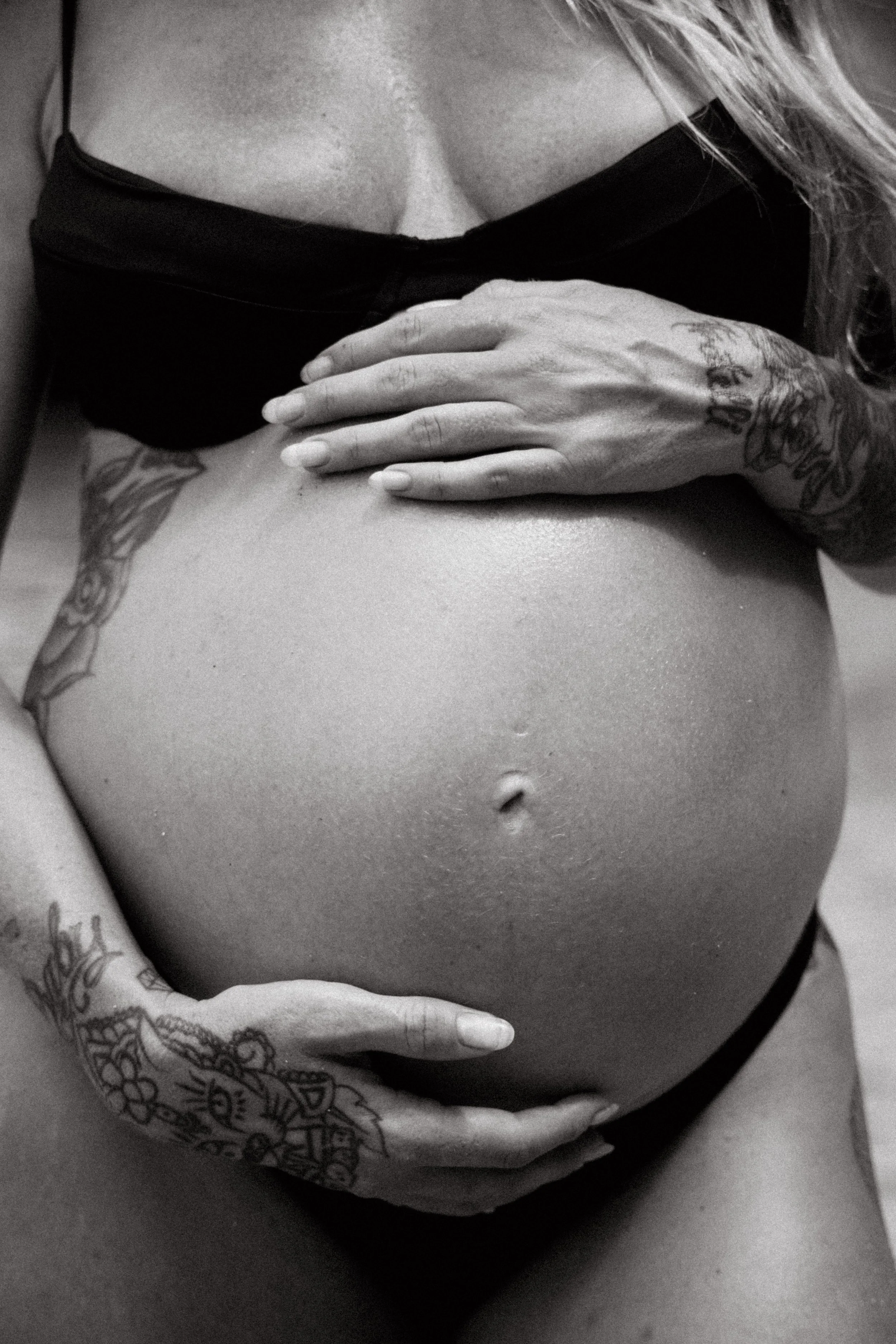 Close-up of a woman's torso with tattoos, one hand holding her breast, wearing a black top, in black and white.