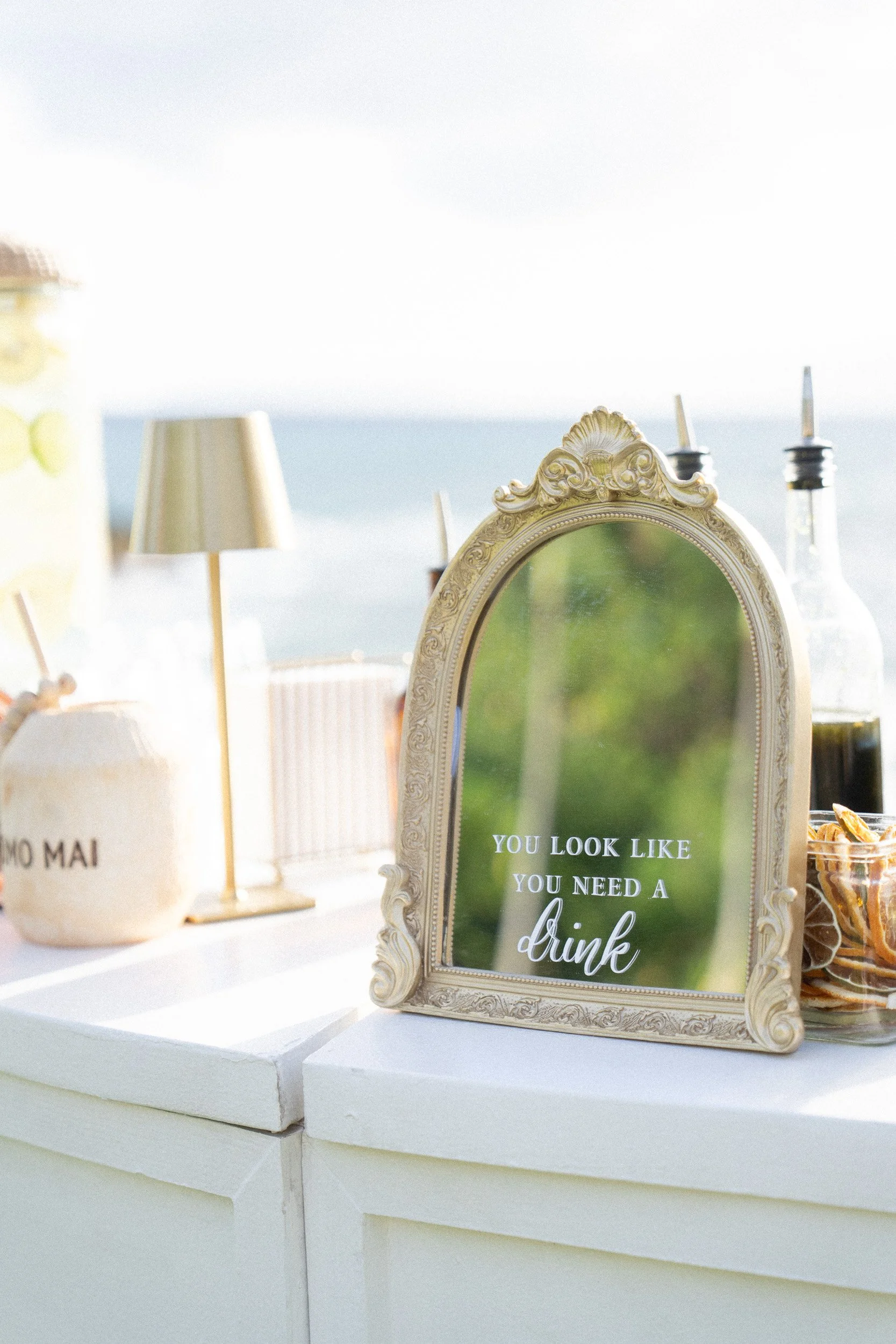 Decorative mirror with the quote, 'You look like you need a drink,' on a green background, displayed on a white table with other decor items including a lamp and bottles, outdoors with a seaside view in the background.
