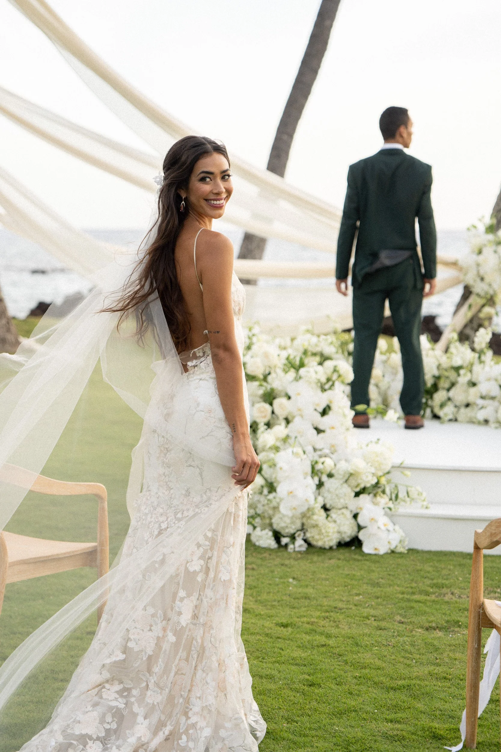 A smiling woman in a wedding dress stands outdoors near a wedding ceremony setup, with a man in a dark suit facing away in the background, and white flowers and greenery around.