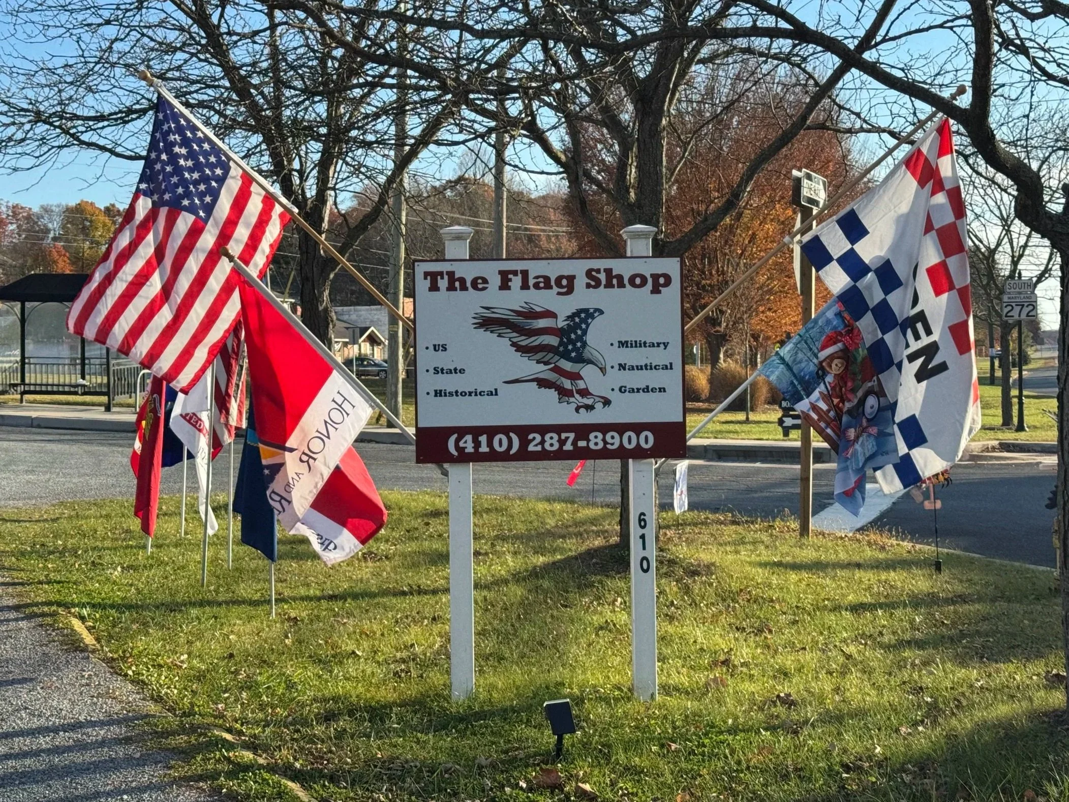 A display of various American flags outside a shop called 'The Flag Shop' with a sign listing services such as US, State, Historical, Military, Nautical, and Garden flags. The shop's contact number is shown. The scene is set in a park-like area with trees and a road in the background, during fall.