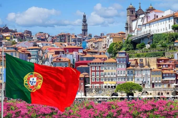 Portugal flag flying in front of a hillside cityscape with colorful buildings and pink flowers at the foreground.