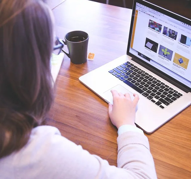Person using a laptop at a wooden desk, with a black coffee mug and an orange tea bag wrapper nearby.