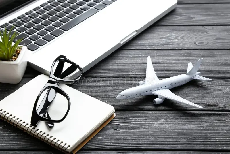 Model airplane, eyeglasses, notebook, and laptop on wooden desk with a potted plant.