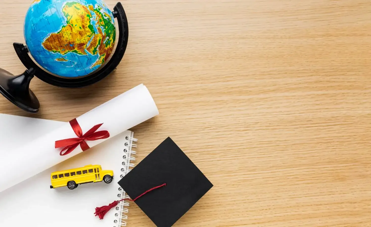 Global globe, rolled white diploma with red ribbon, yellow school bus toy, black graduation cap, and notebook on wooden desk, celebrating education and graduation.
