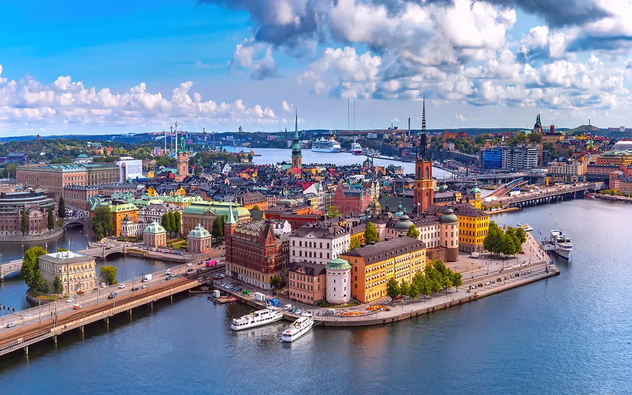 Aerial view of Stockholm, Sweden, with the city skyline, historic buildings, and boats on the water under a partly cloudy sky.