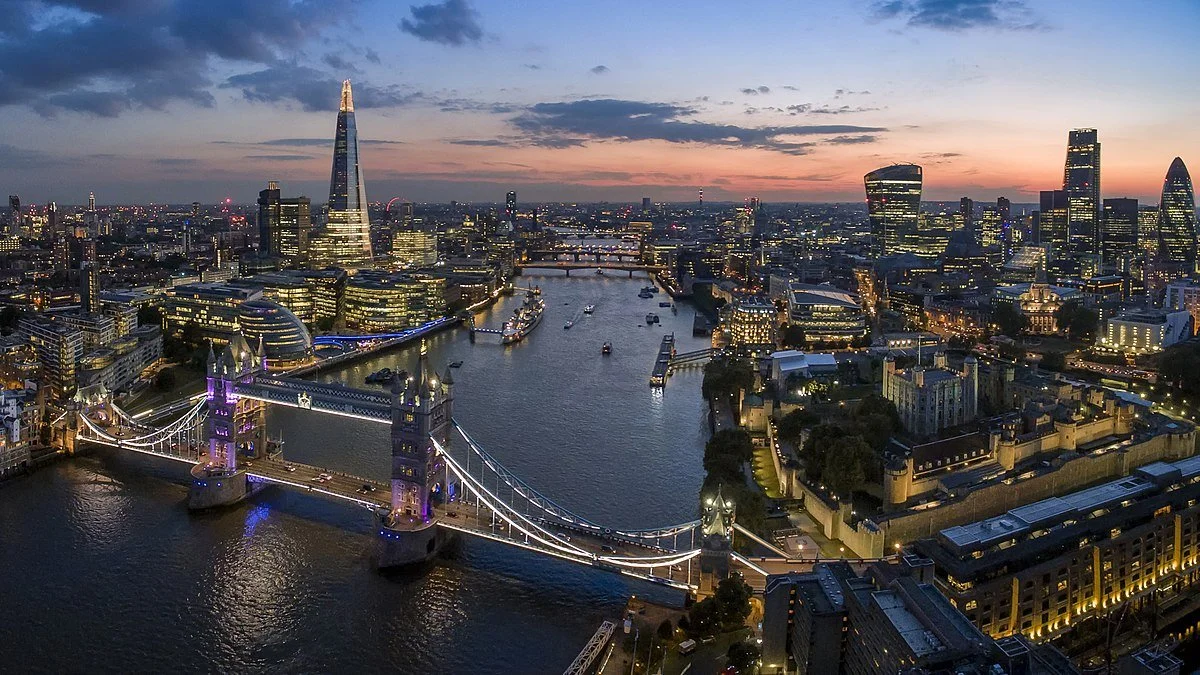 Aerial view of London at sunset displaying Tower Bridge, the River Thames, and the City's skyline with illuminated buildings including The Shard.
