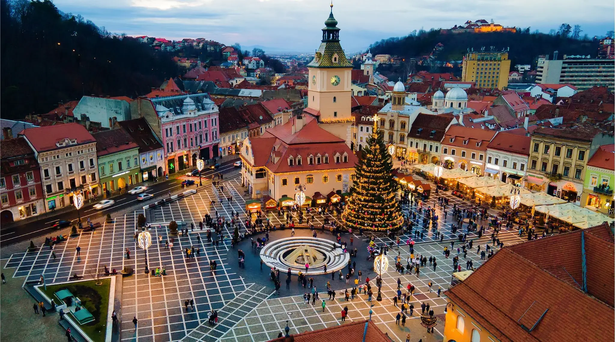 A European town square decorated for Christmas with a large Christmas tree, a fountain, and colorful historic buildings, during evening with lights on.