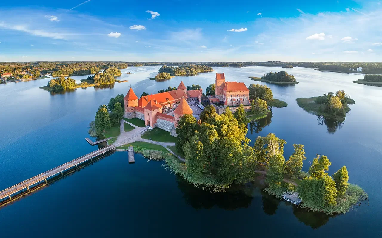 Aerial view of a medieval castle on a small island surrounded by a lake with several smaller islands, trees, and a sky with scattered clouds.