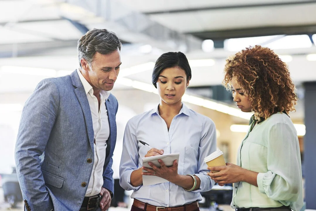 Three diverse professionals having a discussion in an office or workspace, with one person taking notes and another holding a coffee cup.