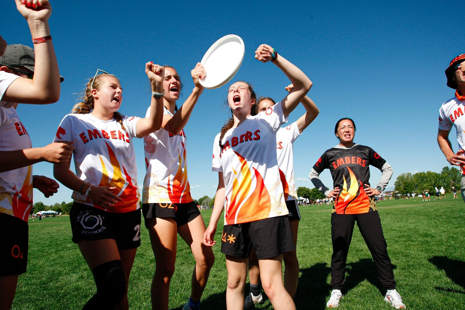Girls in sports uniforms celebrating on a grassy field during a sports event, with blue sky in the background.