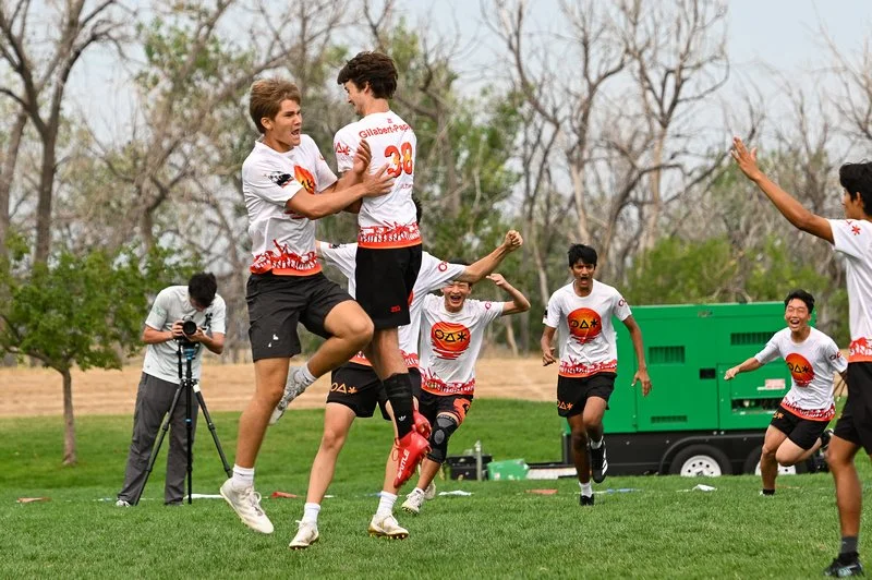 Group of young people on a grassy field, engaging in a sports activity, wearing sportswear with onlookers in the background.