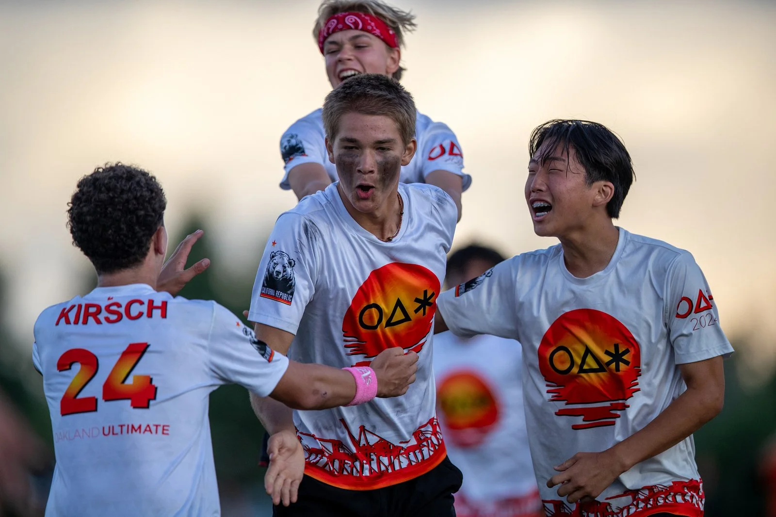 Group of young soccer players celebrating on the field, wearing white jerseys with a red and orange sunset graphic, during daytime.