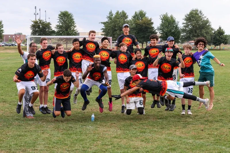 A youth soccer team poses on a field, some kids jumping, others lying or sitting on the grass, with a goalpost and trees in the background.