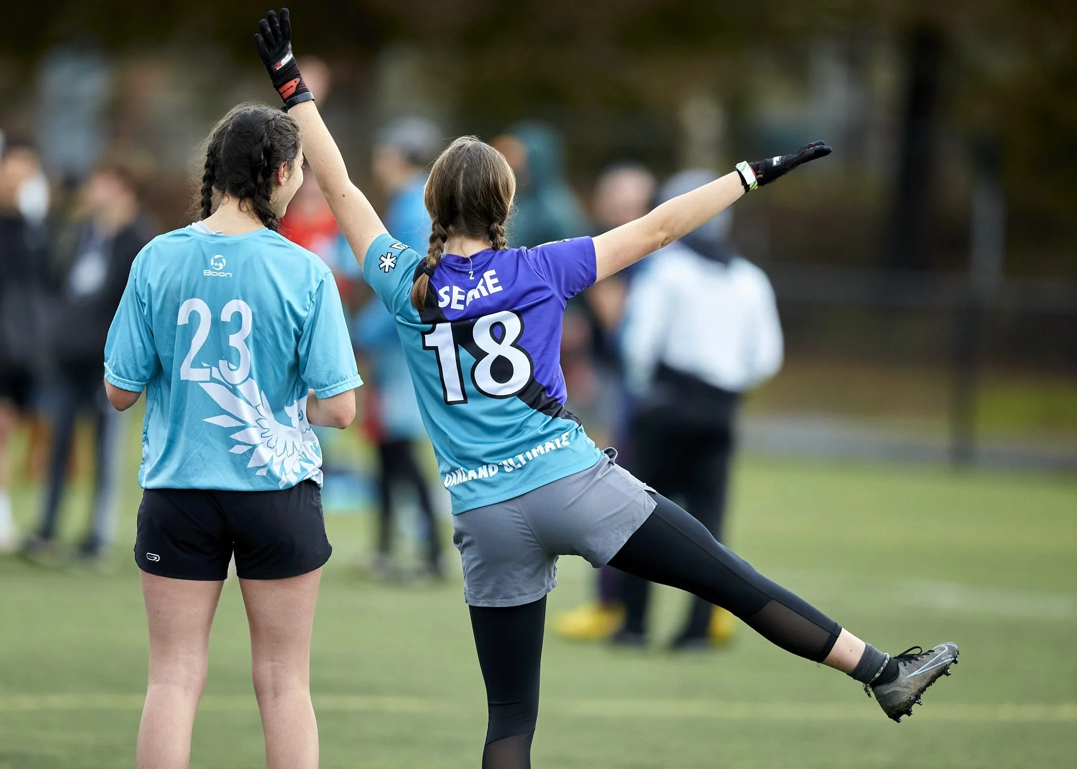 Two female soccer players in blue jerseys with numbers 23 and 18, celebrating on the field. The player with number 18 is kicking one leg up and raising arms, while the other has her back turned. Blurred spectators stand in the background.