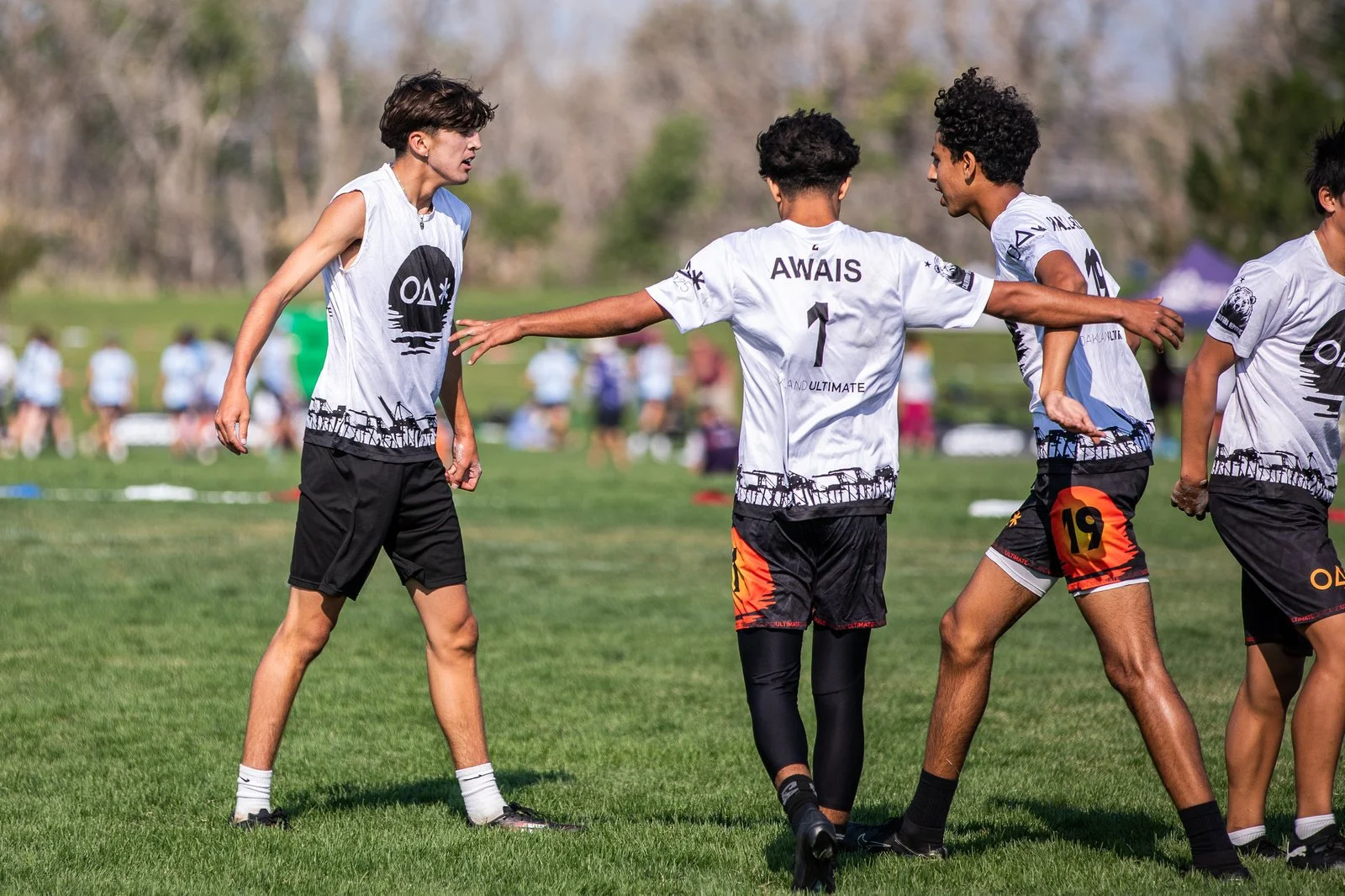 Group of young people on a grassy field, engaging in a sports activity, wearing sportswear with onlookers in the background.