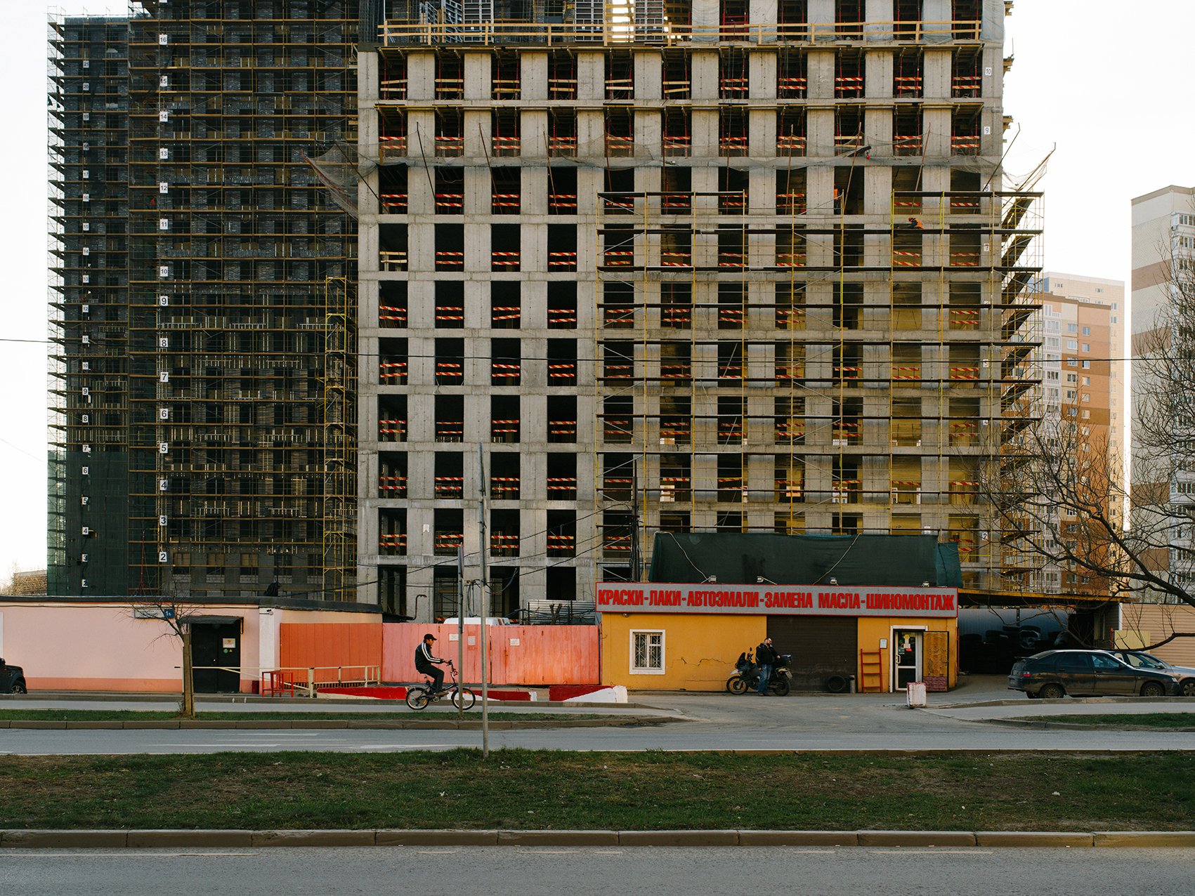 Construction site with a partially built high-rise building and a small yellow shop in the foreground. The shop has a red sign with white text and is located along a street with a cyclist and a motorcyclist passing by.