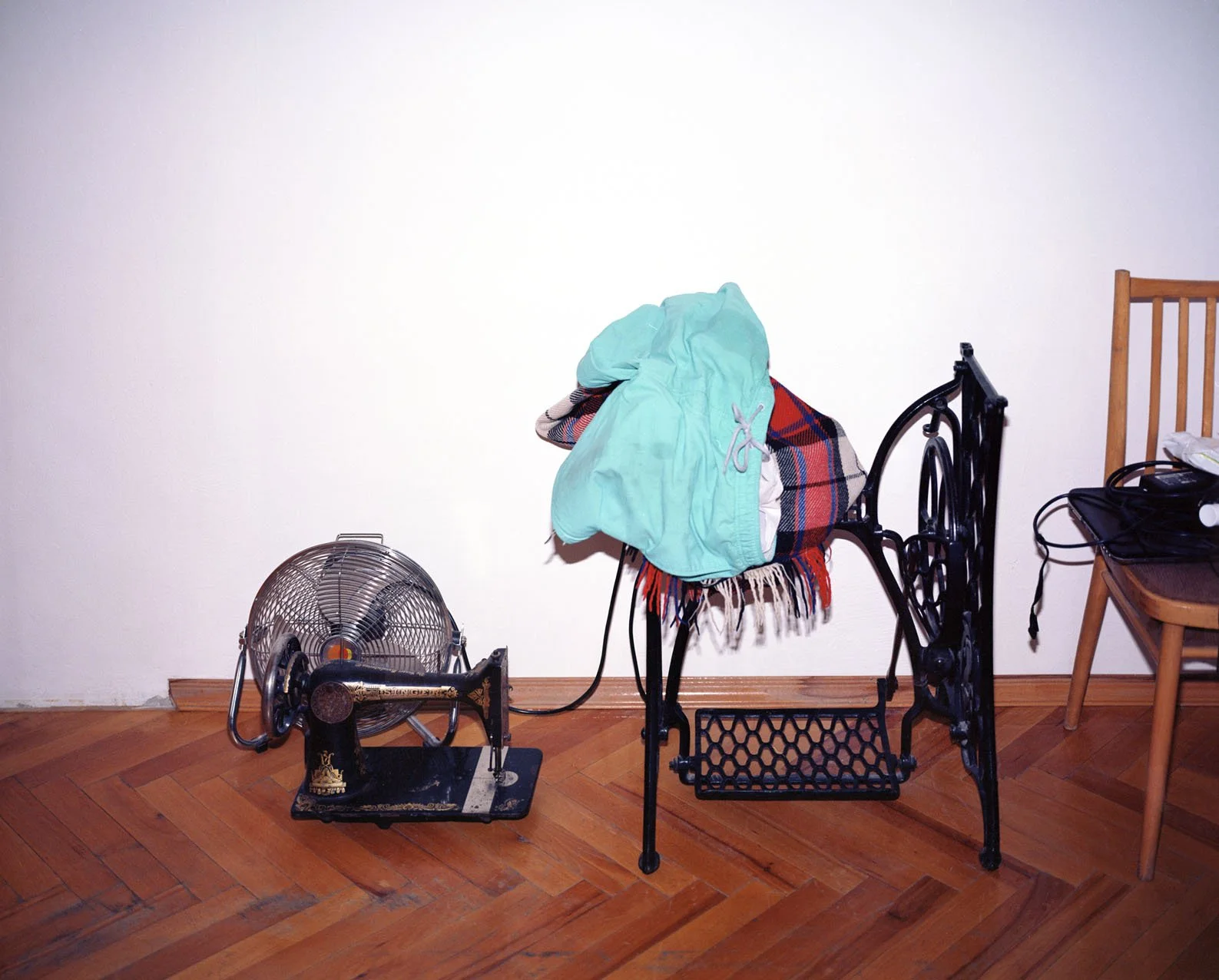 Small table fan next to a black vintage sewing machine with a pile of colorful clothes and blankets on top, and a wooden chair with a black bag on it in a room with wooden floors and white walls.